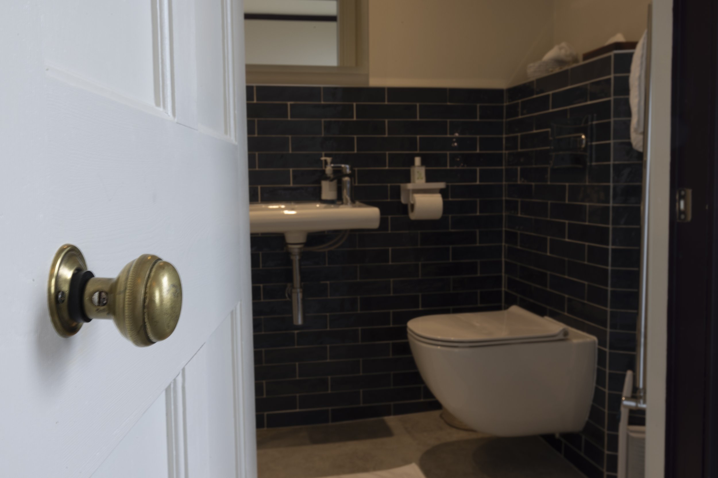 View of a small bathroom with black tiled walls, a white toilet, a small white sink, a soap dispenser, a mirror, a toilet paper holder, and a paper towel dispenser. The door with a brass doorknob is partially open.