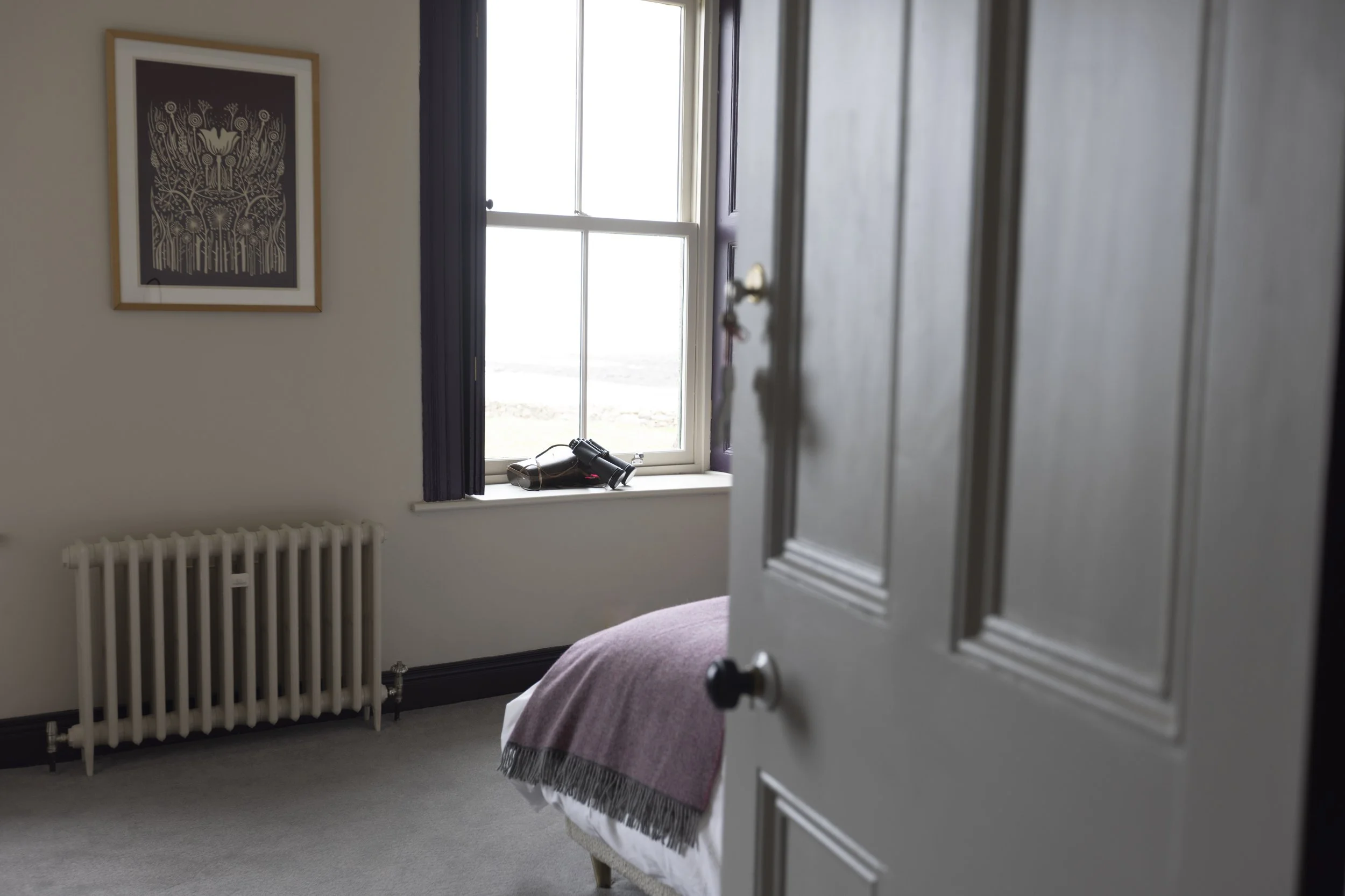 A bedroom with white walls, a framed artwork, a window with dark frames and curtains, a bed with a pink blanket, and a pair of binoculars on the windowsill, seen partially through an open door.