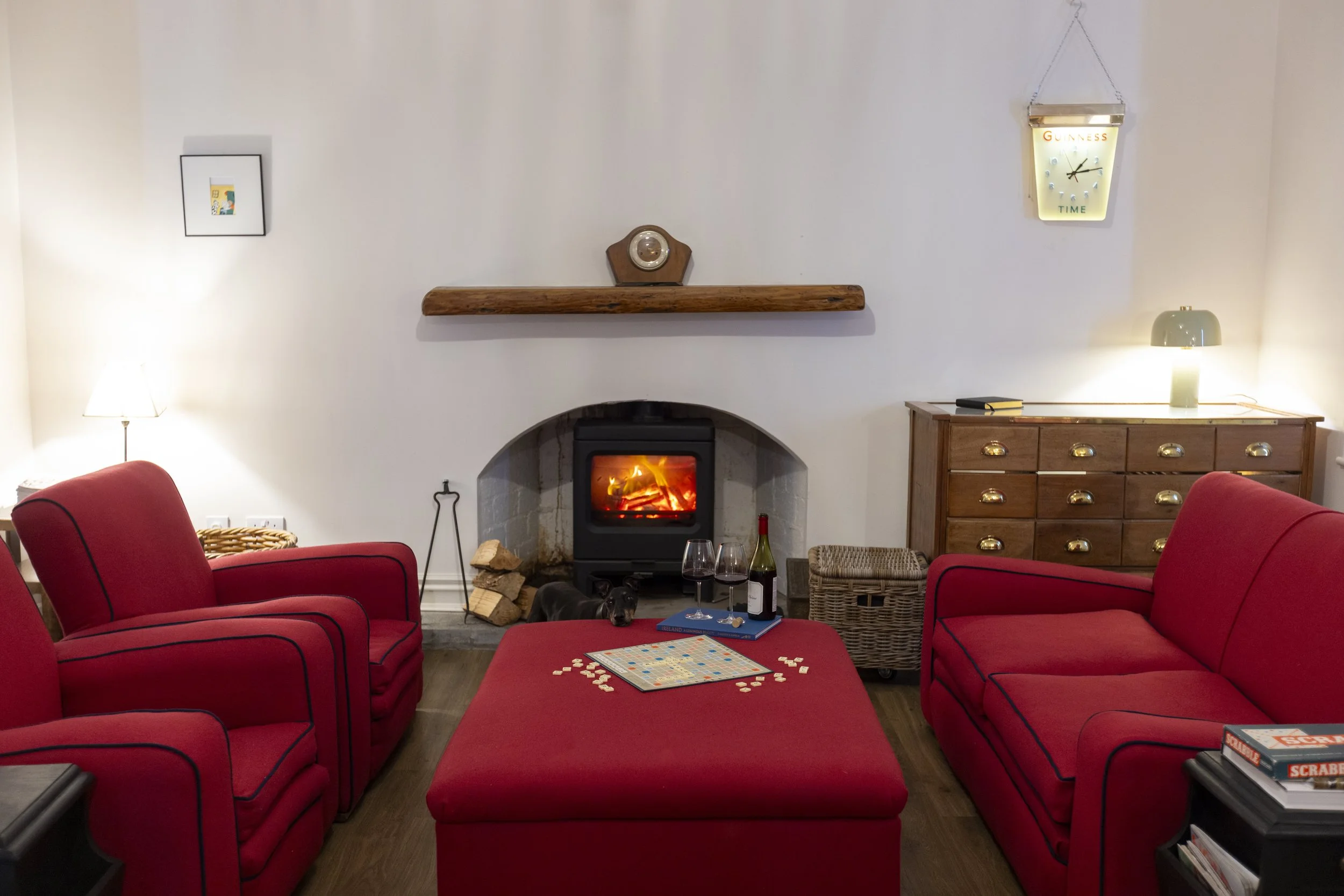 Living room with red upholstered armchairs and a couch arranged around a fireplace with a wood stove. There are glasses of wine, a chessboard, and scattered chess pieces on the ottoman. The room features a wooden cabinet, a small table lamp, a wall clock, and a framed picture on the wall.