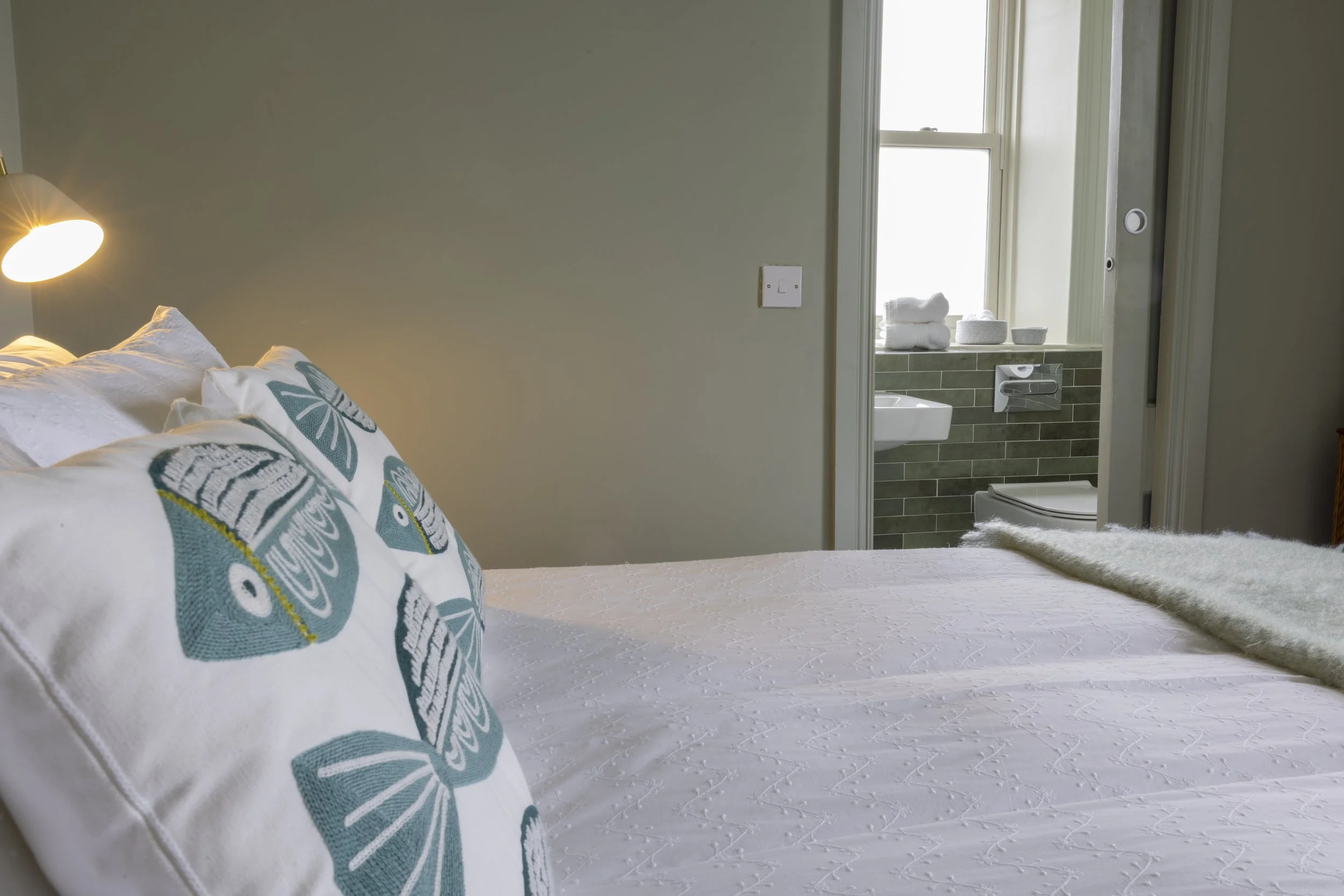 Bedroom with a bed having white linens and decorative pillows, a wall-mounted lamp, and a view into a bathroom with towels and toiletries on the windowsill.