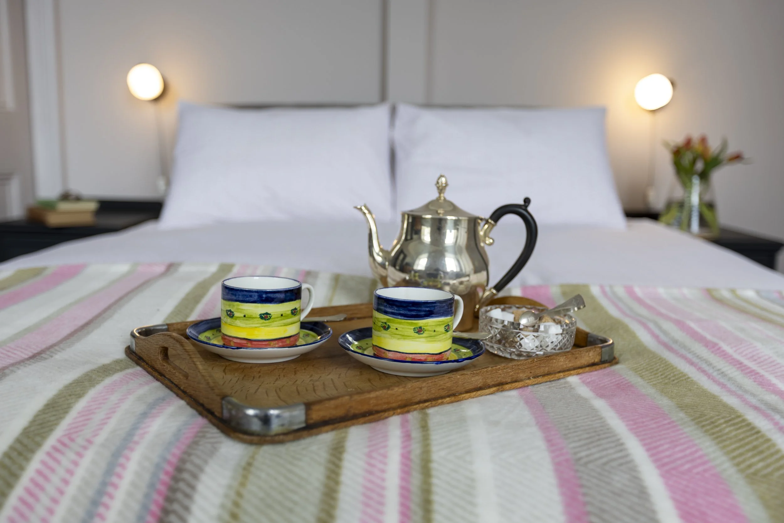 A tray with two colorful coffee cups, a silver teapot, and a glass bowl with sugar packets on a bed with a striped pink, green, and cream bedspread in a bedroom with bedside lamps and a flower vase.
