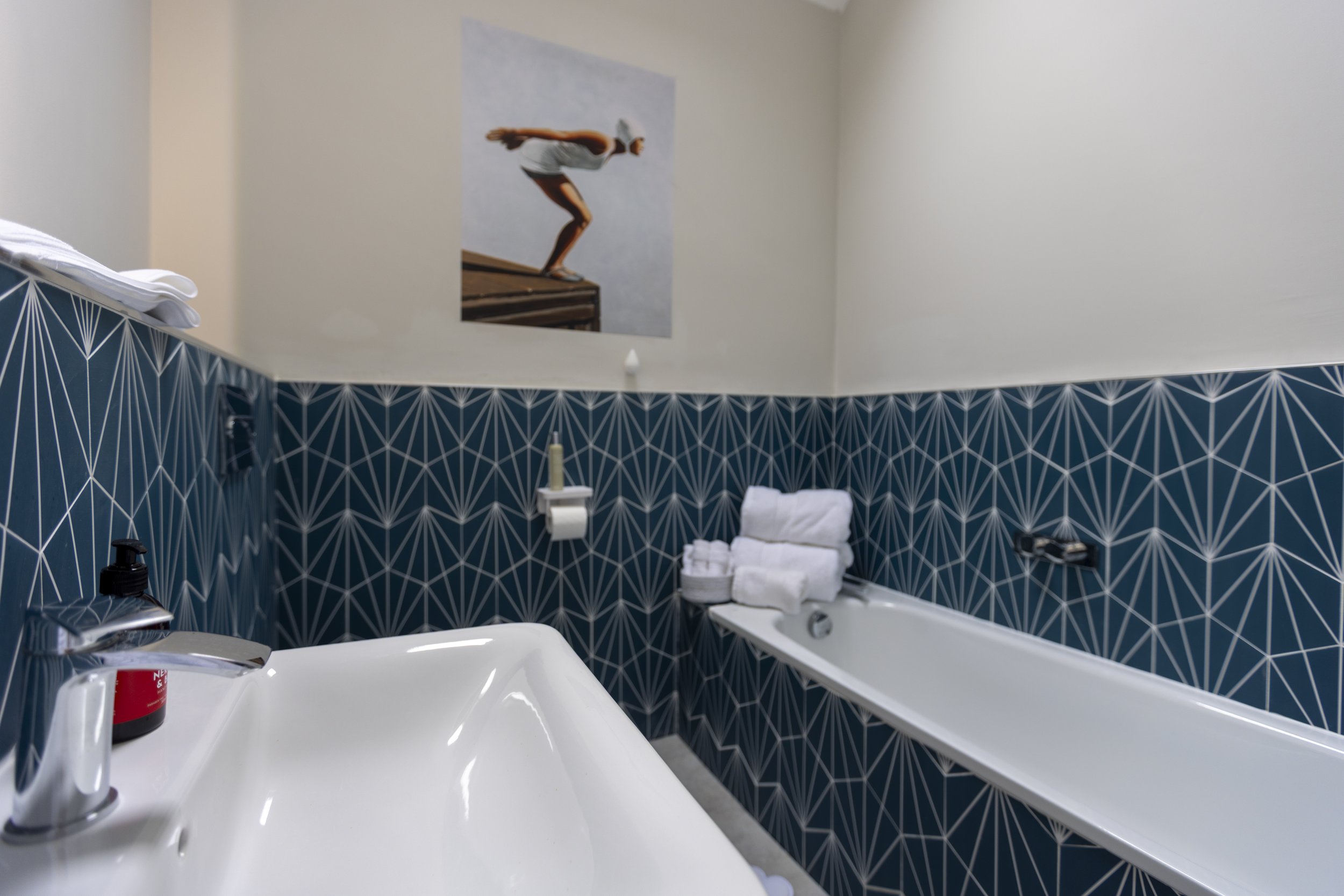 Bathroom with a patterned blue tile wall, a bathtub with white towel folded on the edge, a sink with soap, a wall-mounted toilet paper holder, and a large framed photo of a swimmer on the wall.
