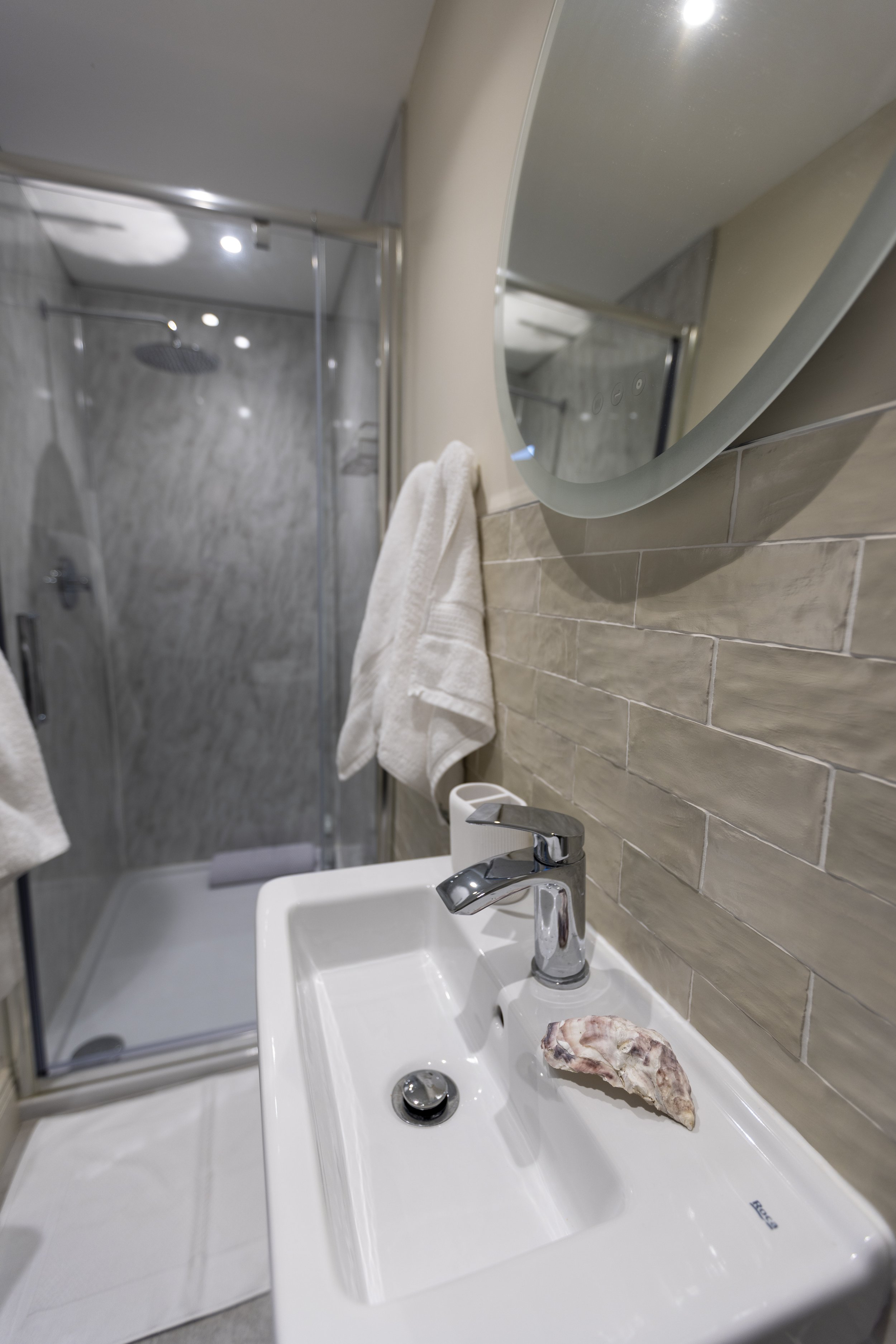 A small bathroom with a white sink, a mirror, and a shower with a glass door. The sink has a piece of rock on it. Towels are hanging on the wall, and the shower area has marble-like tiles.