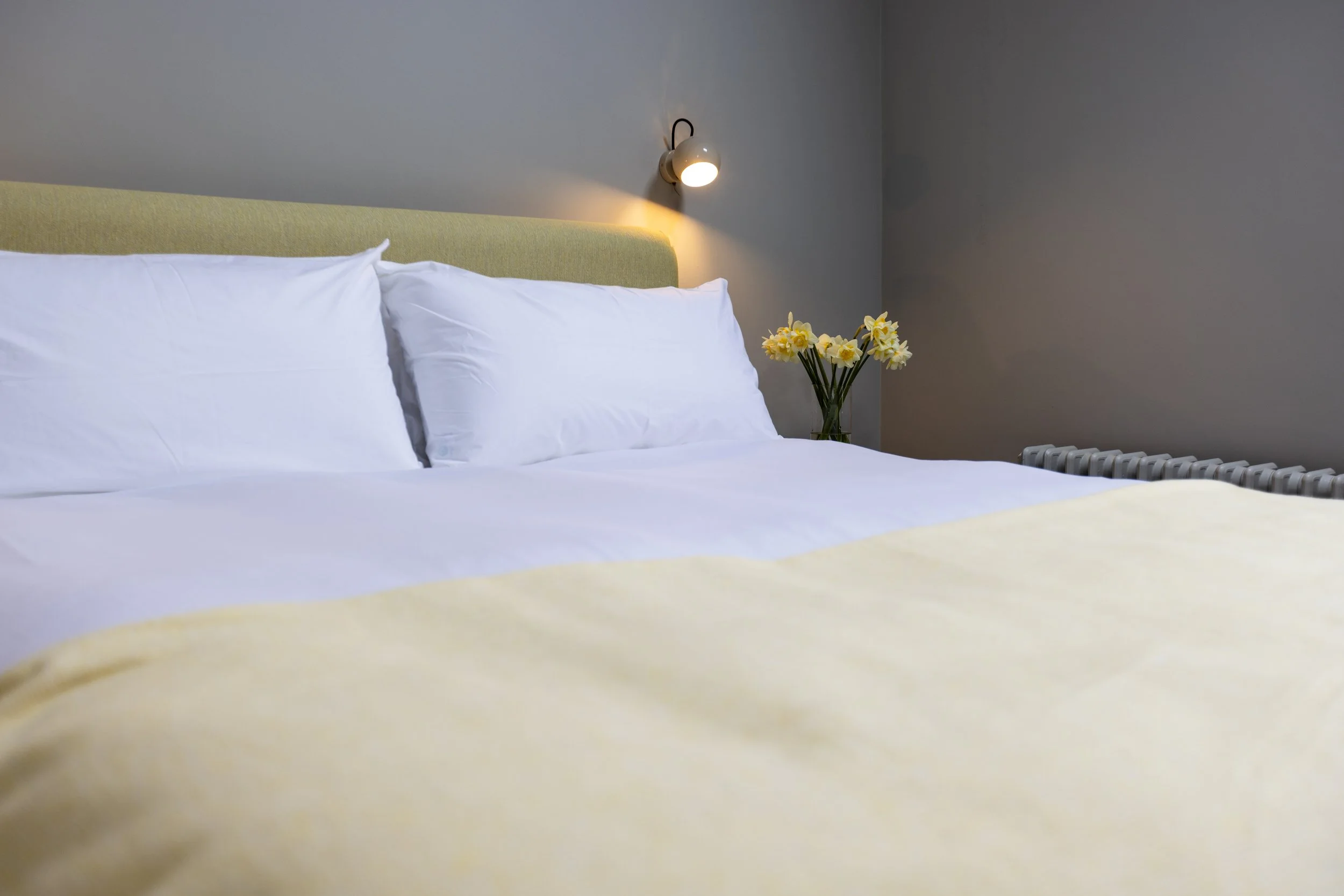 A neatly made bed with white sheets and pillows, a yellow blanket, a yellow upholstered headboard, a wall-mounted reading lamp, a vase with yellow and white flowers, and a radiator beside the bed.