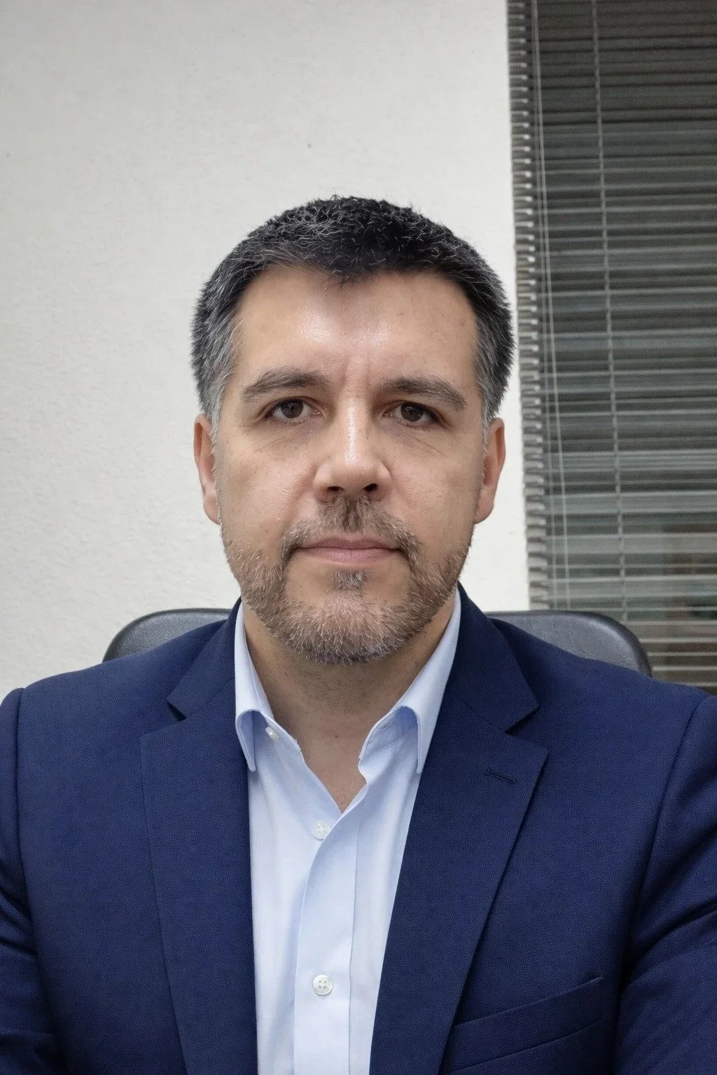 Retrato de un hombre con barba y cabello oscuro, vestido con Americana azul y camisa blanca, sentado en una oficina con pared blanca y persiana de ventanas en el fondo.