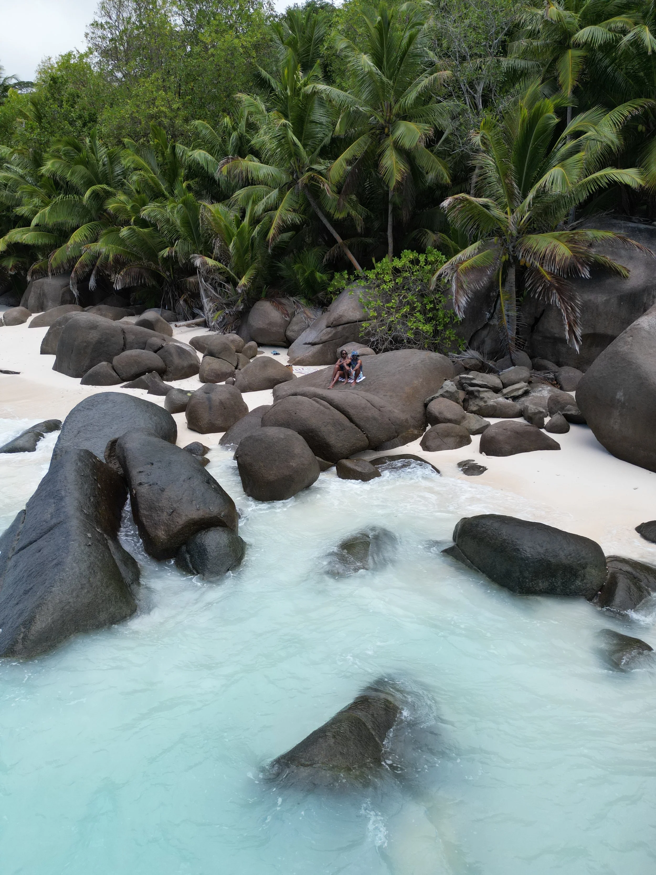 Tropical beach with white sand, large rocks, and palm trees in the background; two people sitting on a large rock near the shoreline.