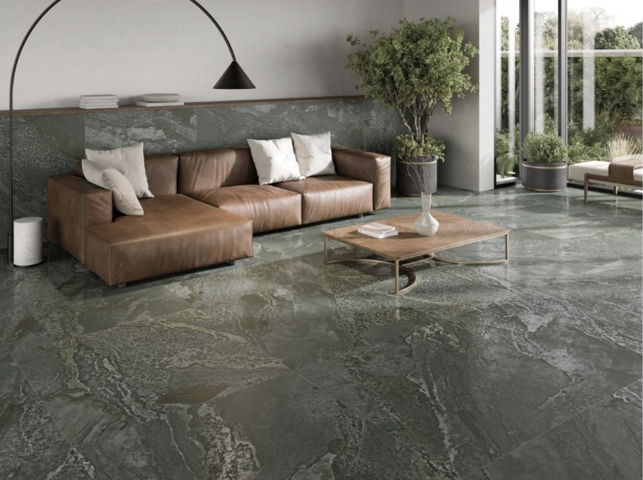 Modern living room with brown leather sectional sofa, white pillows, a wooden coffee table with a white vase and books, large windows with greenery outside, potted plants, and minimalistic decor.