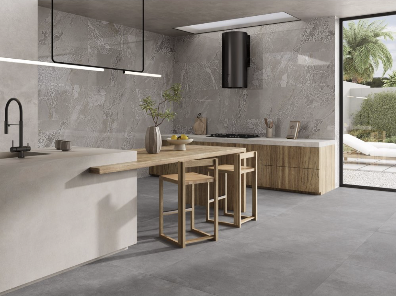 Modern kitchen with gray tile floors and walls, a black range hood, light wood cabinets, and a large glass sliding door overlooking a garden with palm trees.