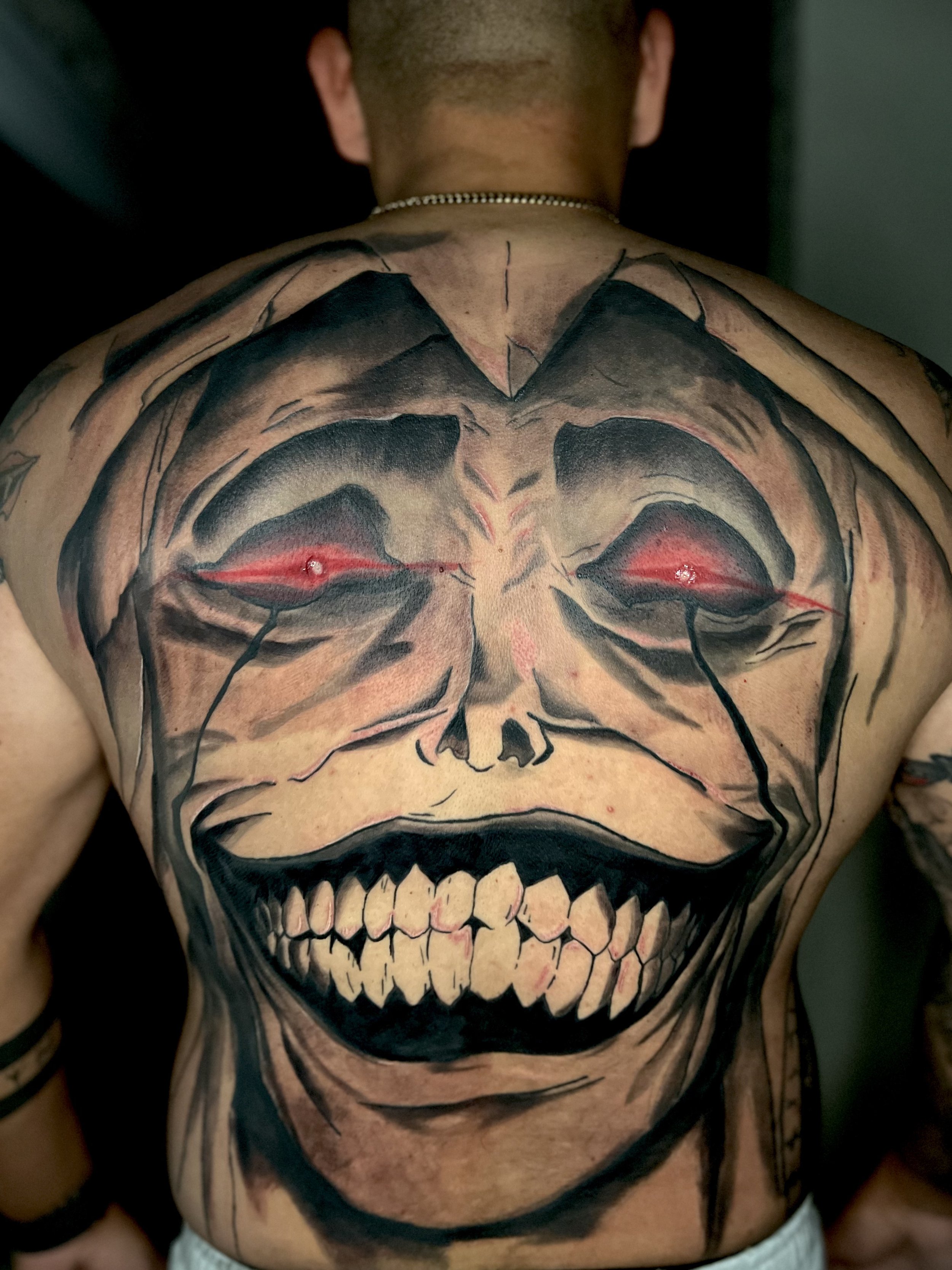 A large, colorful tattoo of the statue of god from the anime solo leveling with glowing red eyes, sharp teeth, and a wide grin covering the person's back.