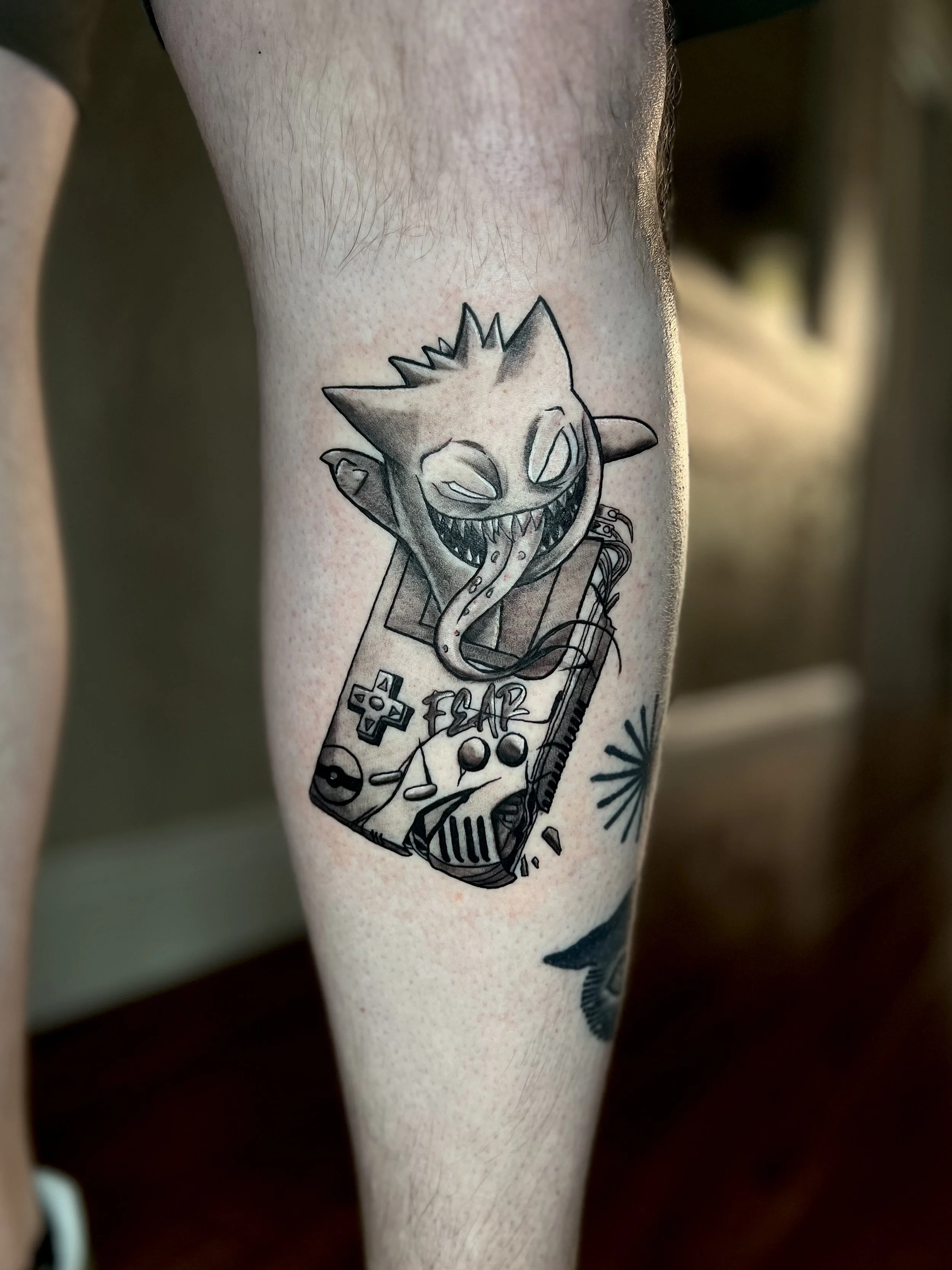 Black and gray tattoo of a sinister, smiling creature with horns and a long tongue, emerging from a vintage gaming controller with various buttons, a plus sign, a yin-yang symbol, and wires.