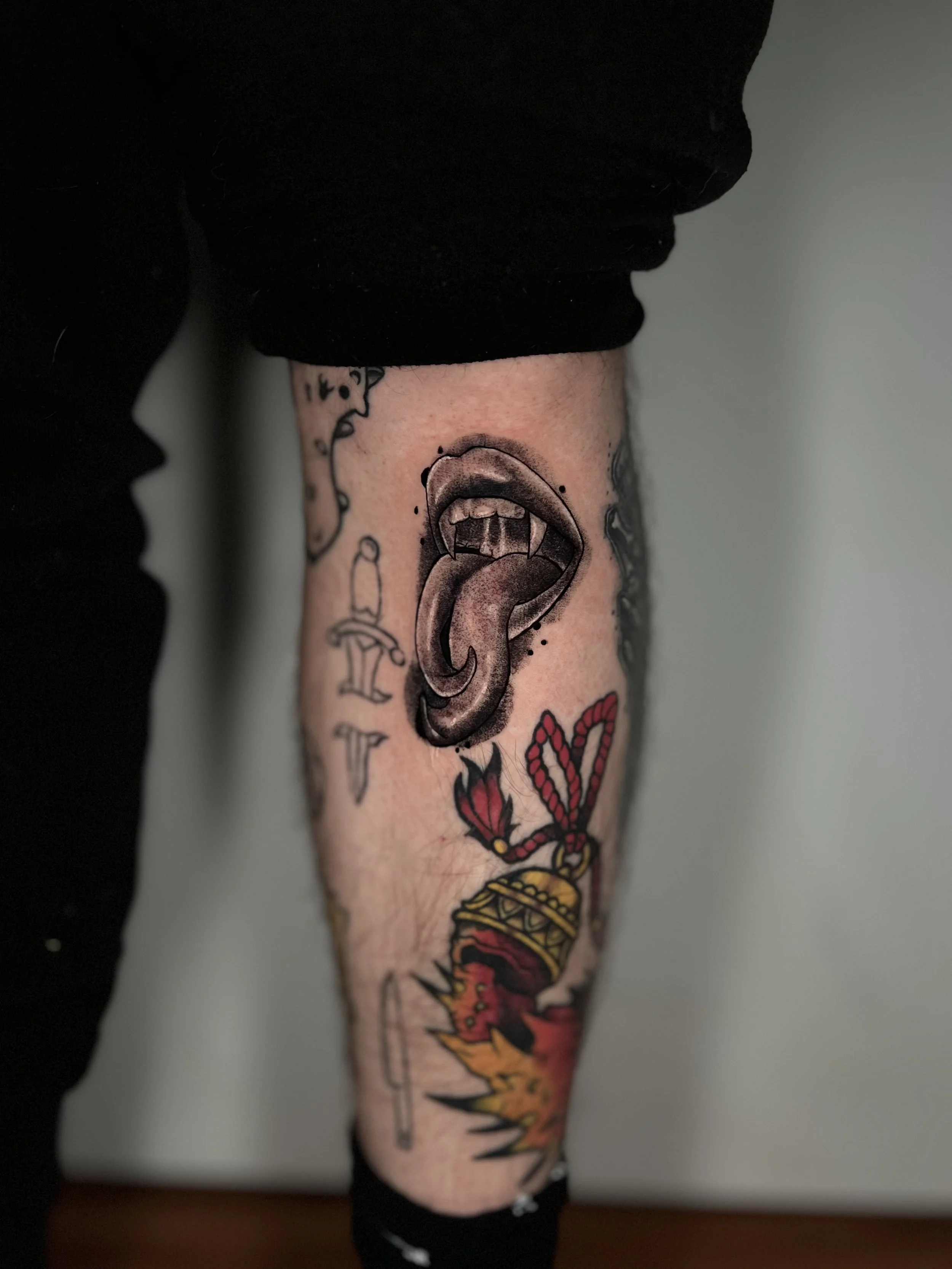 Tattoo of a wide-open mouth with a tongue sticking out on a person's lower leg, with additional tattoos of knives, a heart with a rope, and a burning explosion.