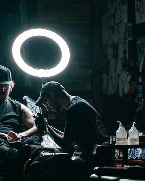 Tattoo artist working on a client's arm in a tattoo studio, illuminated by a large ring light