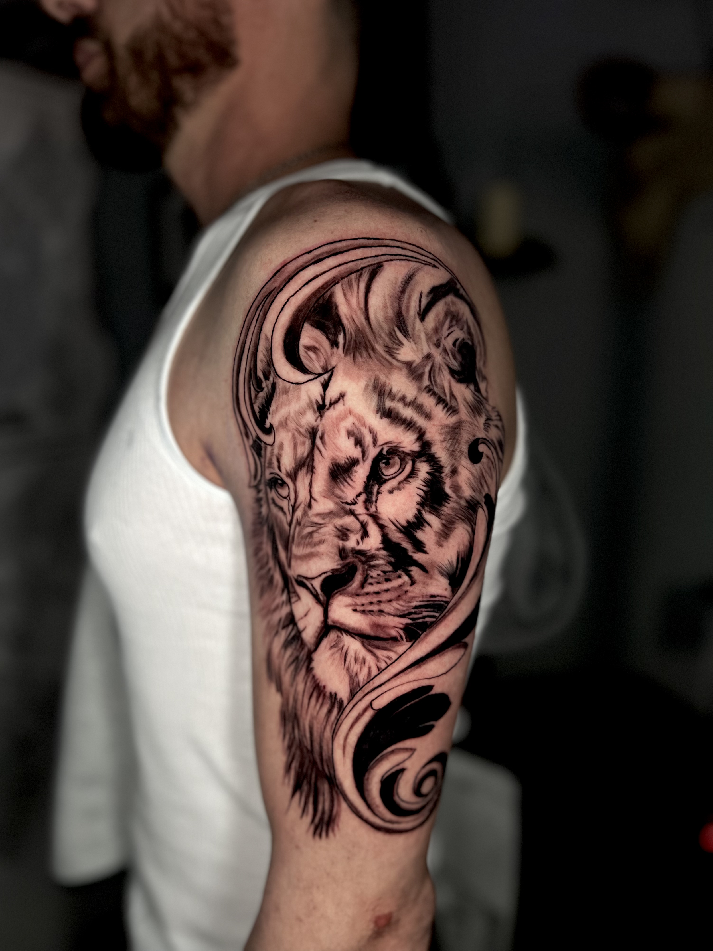 Black and gray tattoo of a lion's face on a man's upper arm, with swirling decorative elements.