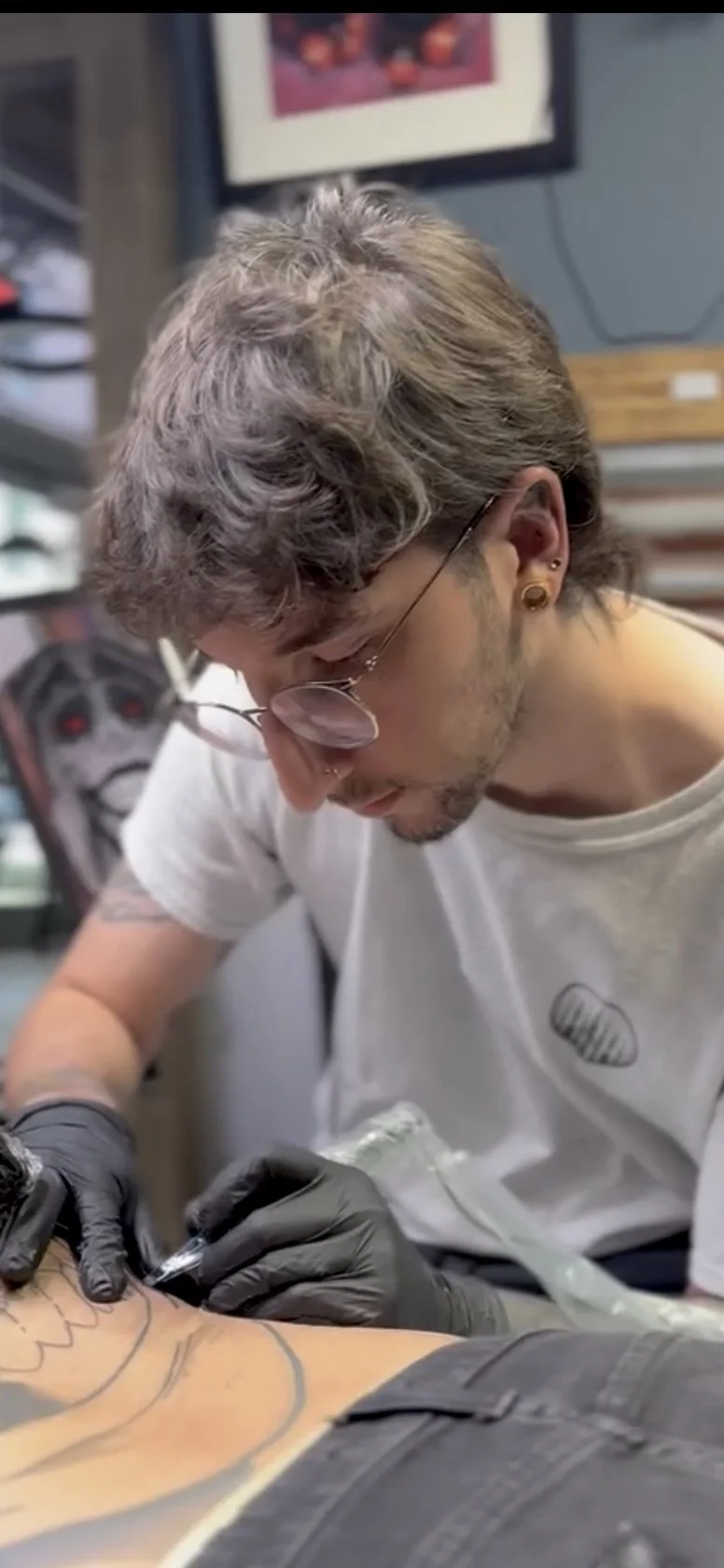 A tattoo artist with glasses and gauged ears creates a tattoo on a client in a tattoo shop.
