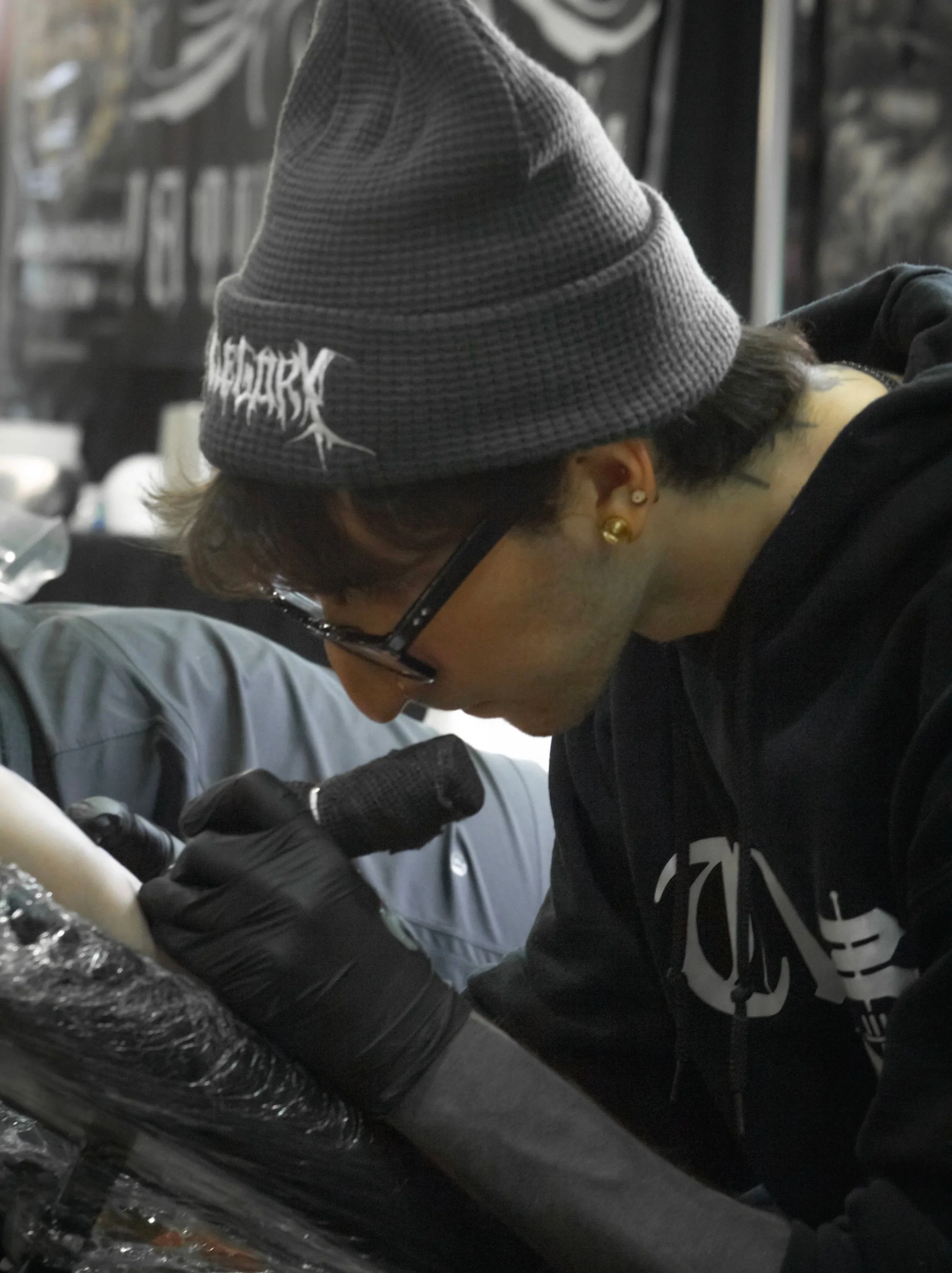 Tattoo artist with glasses, gray beanie, black hoodie, and earrings tattooing a client's arm in a tattoo studio.