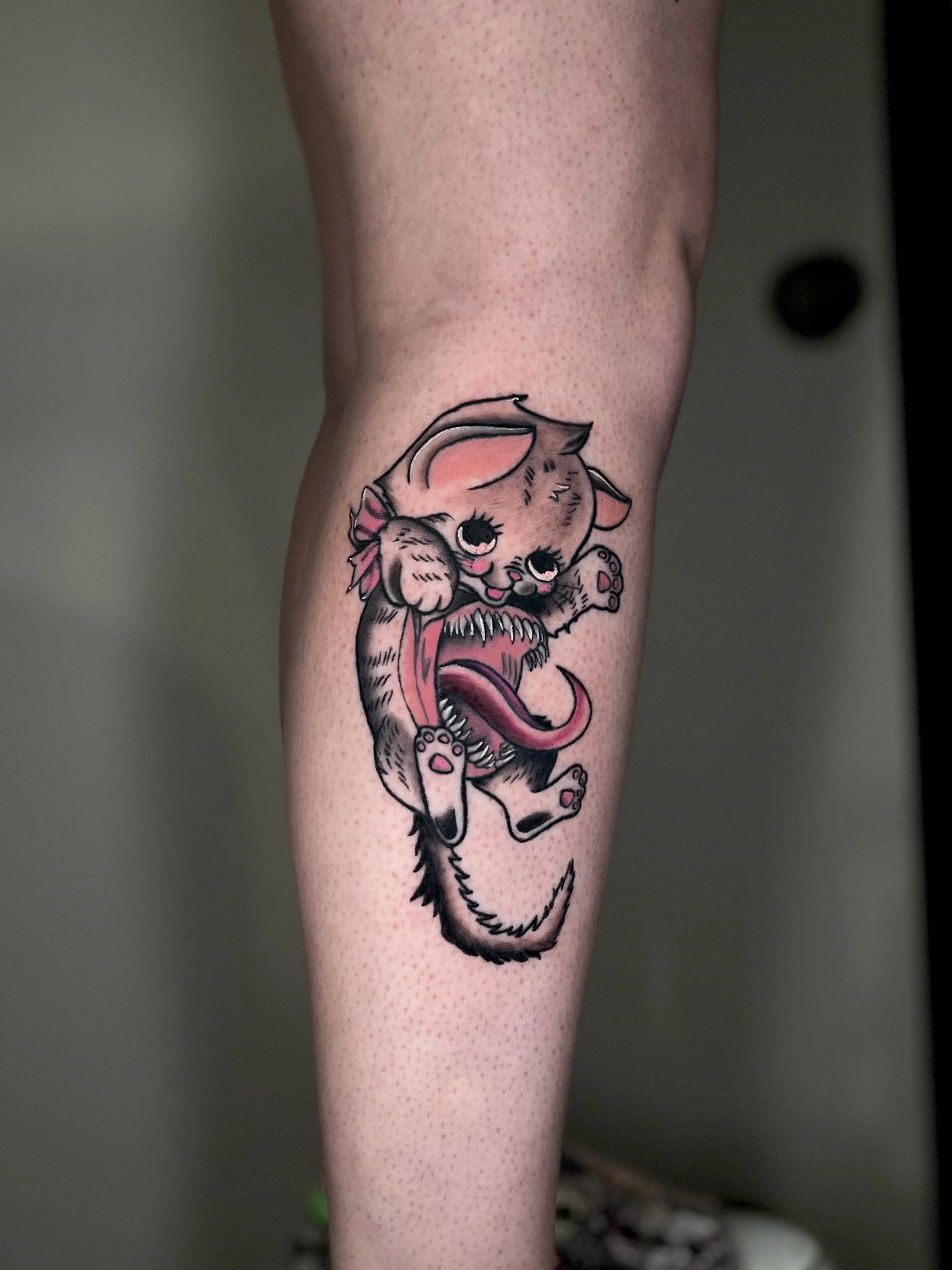 Tattoo of a cute, cartoon-style puppy with large eyes, pink accents, and an exaggerated open mouth showing sharp teeth inside, sitting on a person's leg.