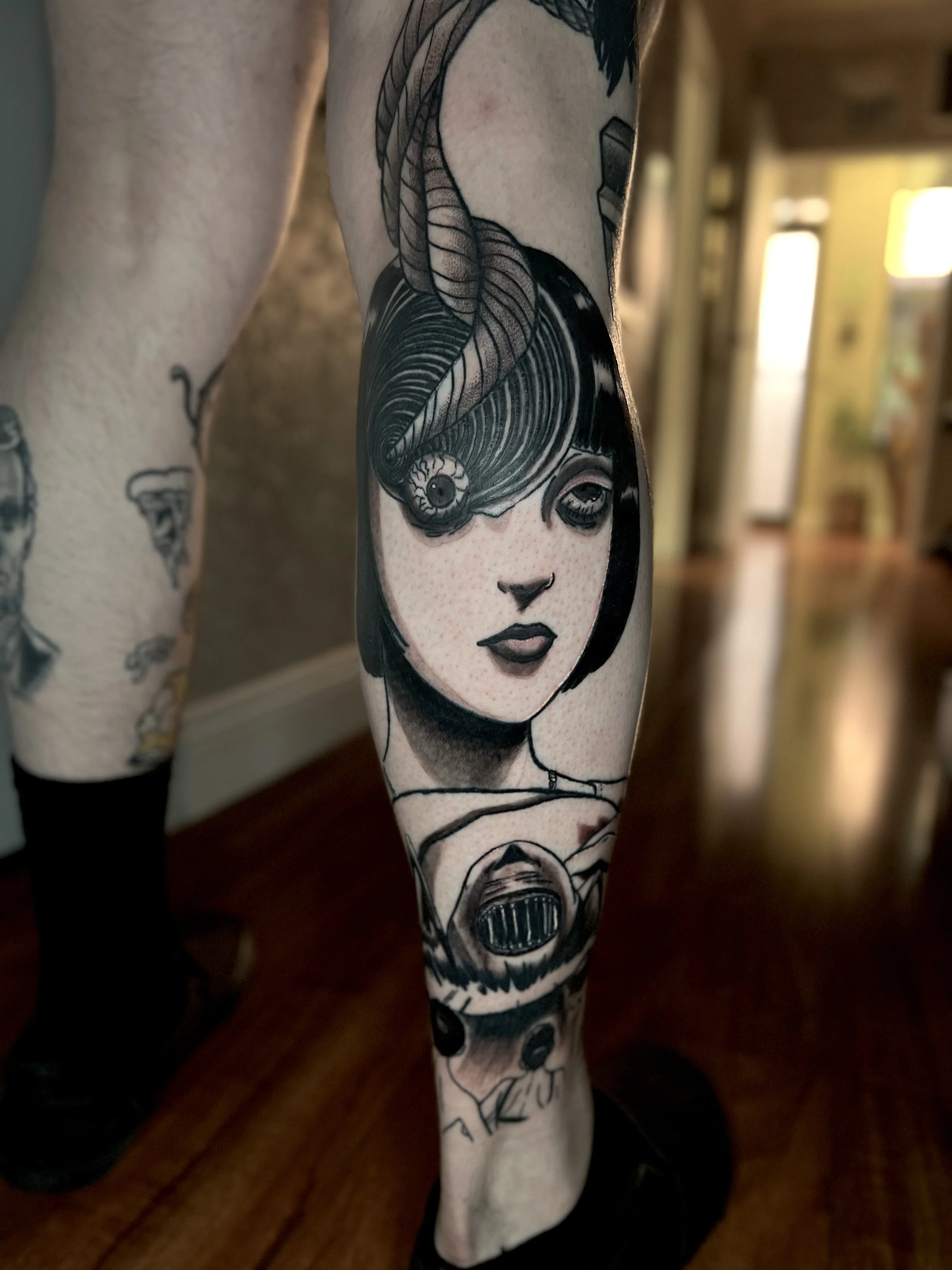 Tattoo of a woman's face on leg with a stylized eye covering one eye, and a robotic skull below, in black and gray ink.