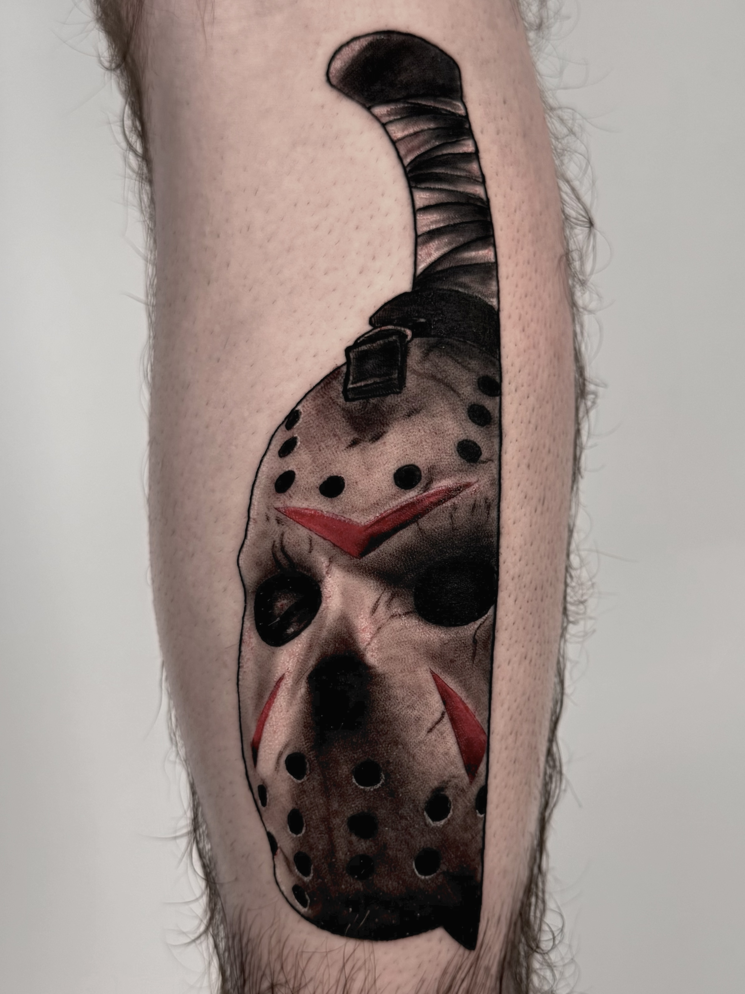 Tattoo of a hockey mask with red markings and black eye holes, resembling Jason Voorhees from horror movies, on a person's arm.