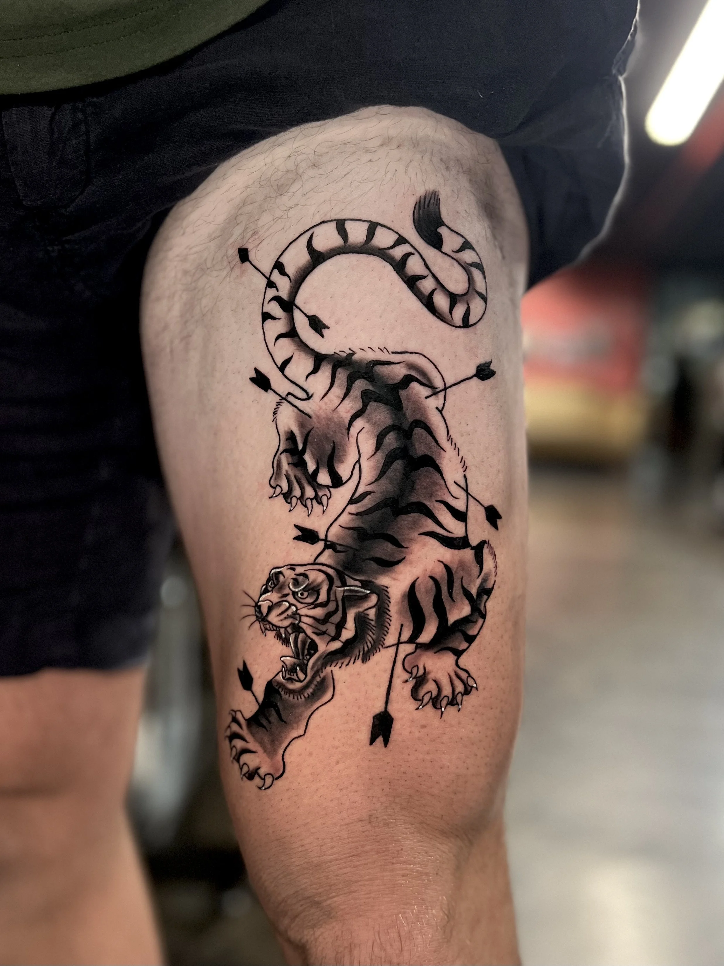 Black and gray tattoo of a fierce tiger with an archer's bow and arrows, depicted in a dynamic, aggressive pose on a person's upper thigh.