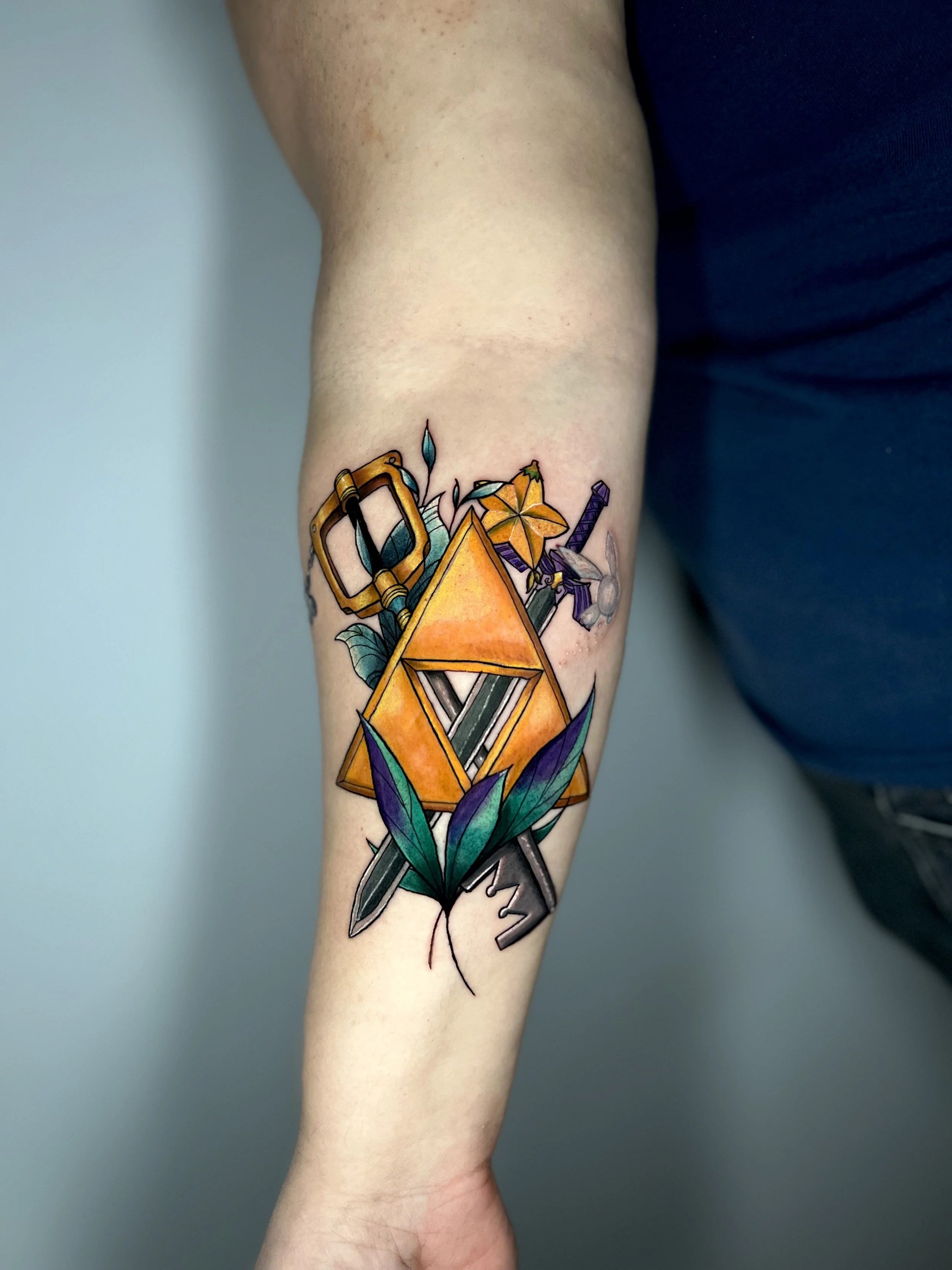 Colorful tattoo on forearm featuring a Triforce from legend of zelda, a key blade from kingdom hearts. inside an orange triangle, swords, leaves, a star-shaped flower, 