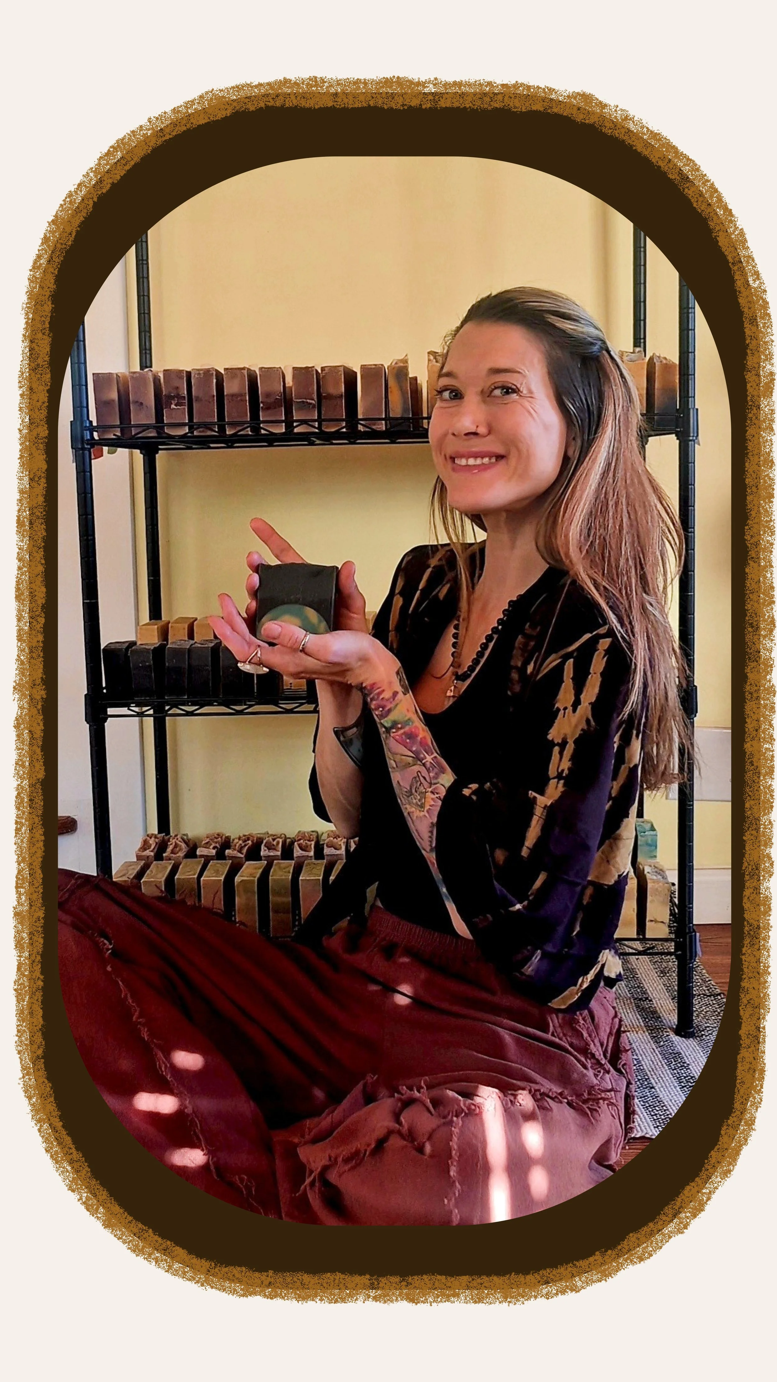 A woman with long hair and tattoos on her arm, smiling and sitting cross-legged, holding soap that has the earth inside of it. She is indoors, with shelves of soap that she made