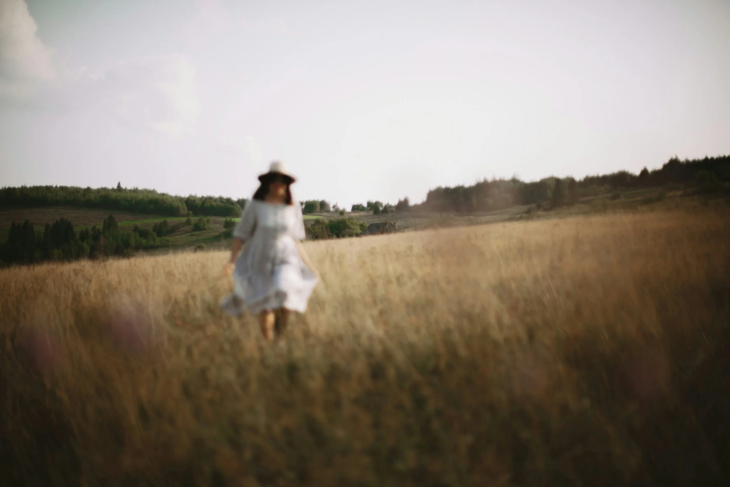 A woman in a white dress and wide-brimmed hat walking through a golden field under a partly cloudy sky, with trees and hills in the background.