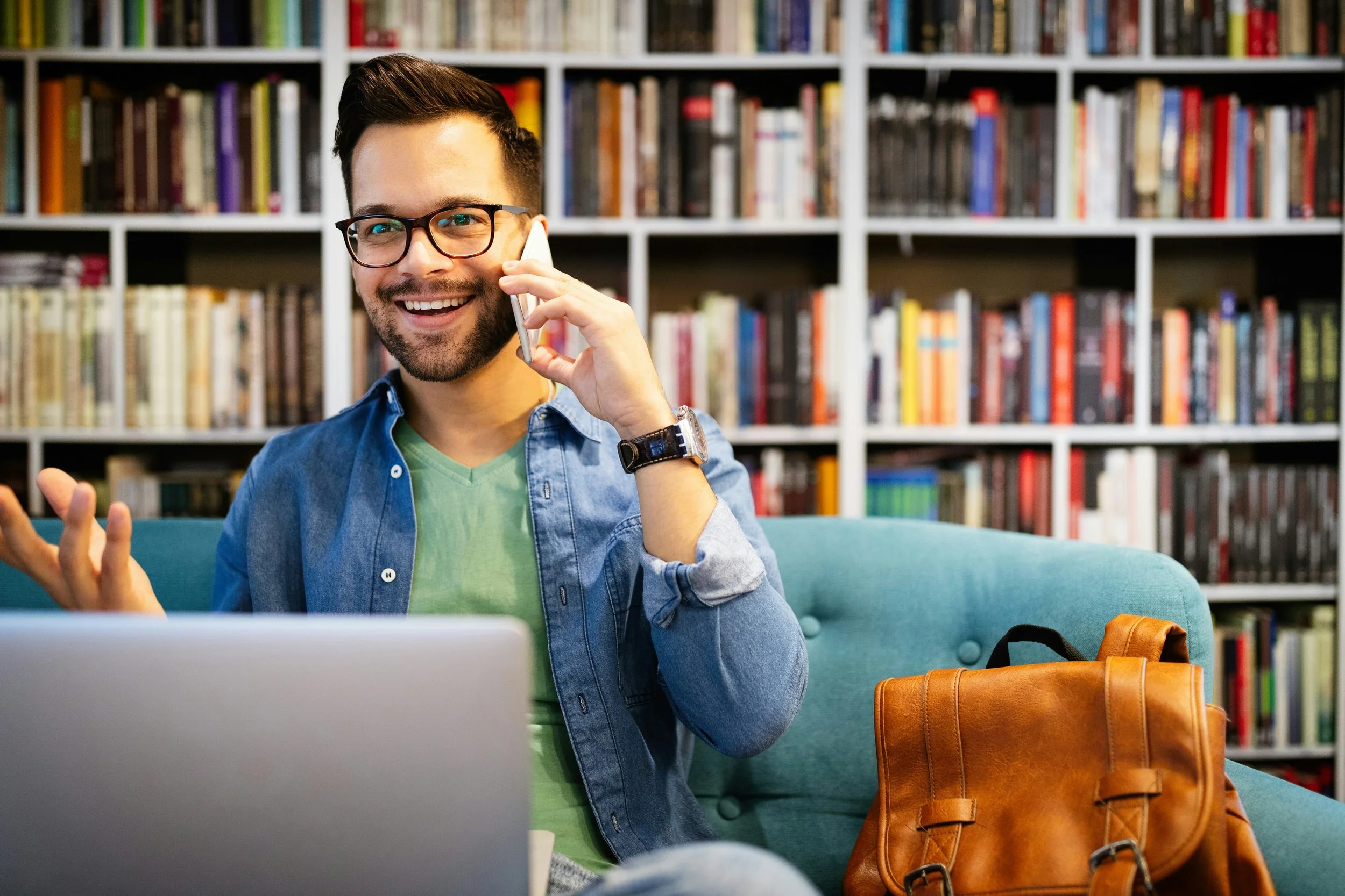 A young man with glasses, a beard, and a smile, talking on a mobile phone while sitting on a teal sofa in a library or bookstore with bookshelves in the background. There is a laptop in front of him and a brown leather bag beside him.