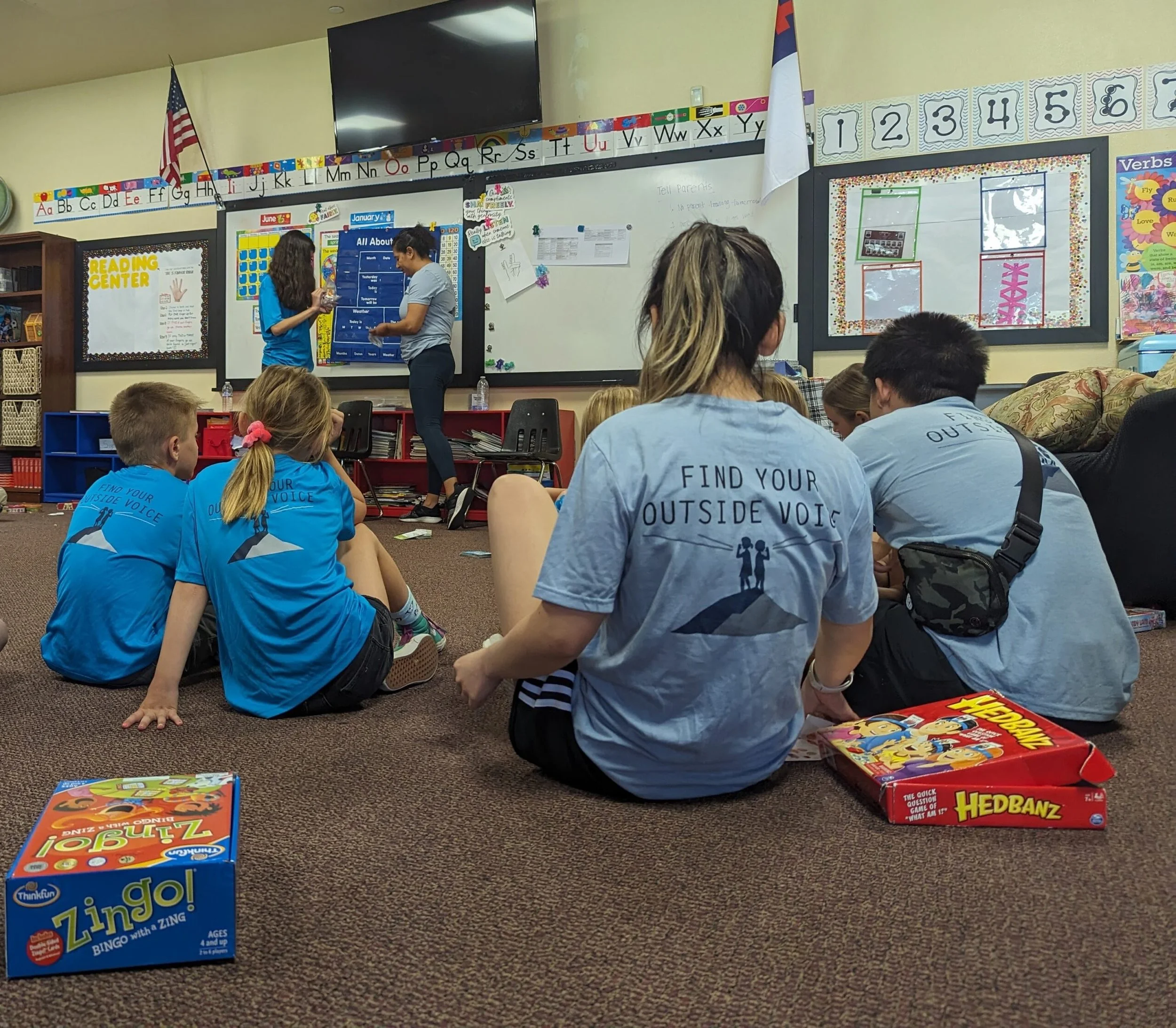 Classroom scene with students sitting on the carpet, some wearing matching blue shirts with the slogan 'Find your outside voice.' Two students stand at the whiteboard, engaged in a presentation, while other students watch. Educational posters and a large TV screen are visible on the wall.