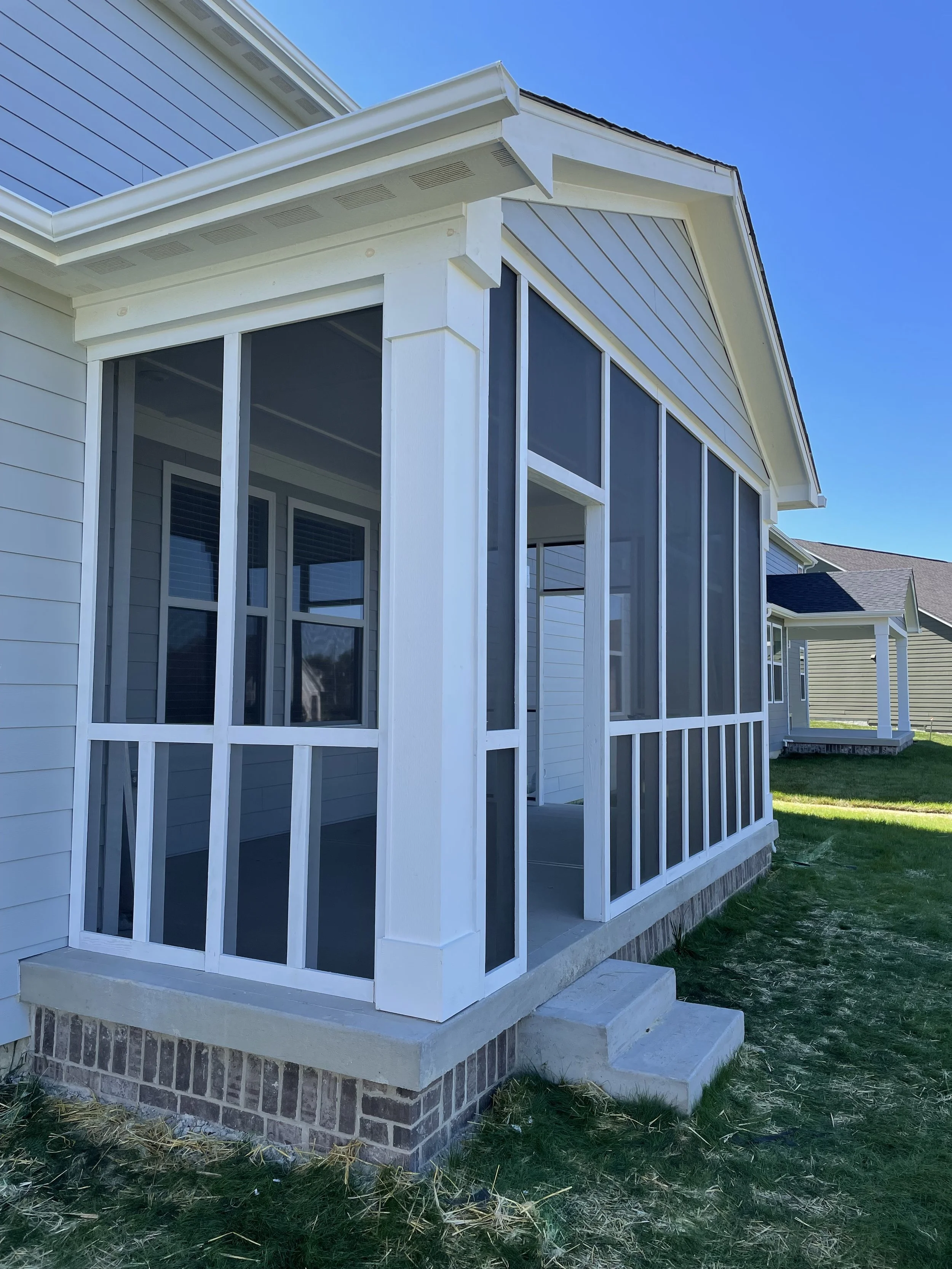 Newly constructed screened porch with white framing, glass panels, and brick foundation attached to a house under a clear blue sky.
