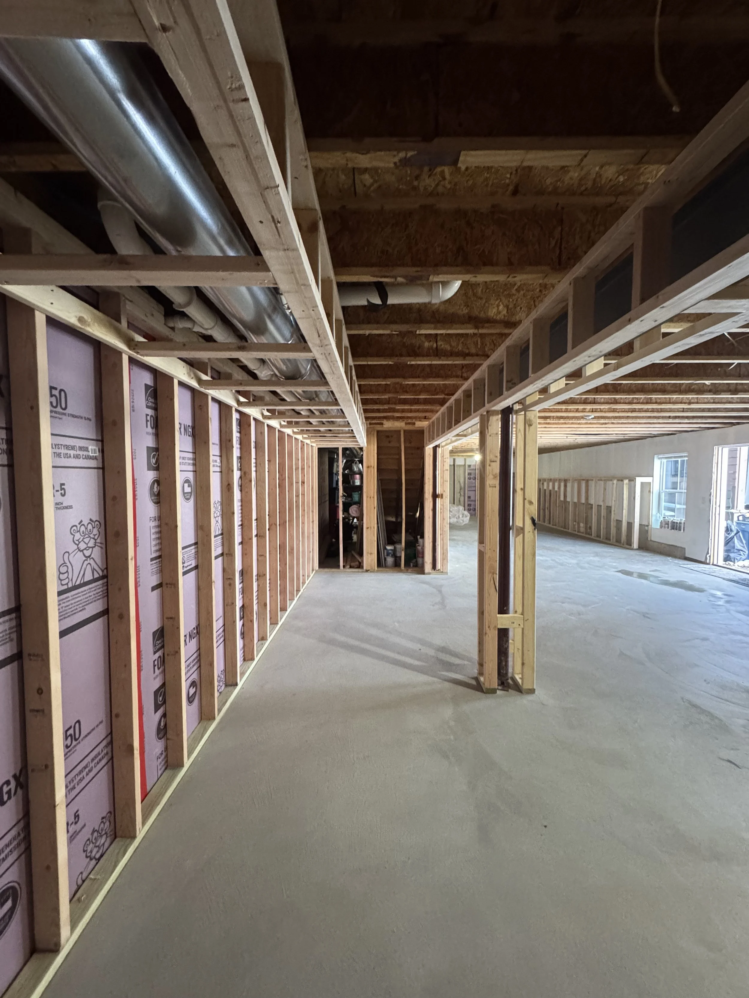 Interior of a house under construction, showing exposed wooden framing, insulation, and ductwork, with a concrete floor and open space.