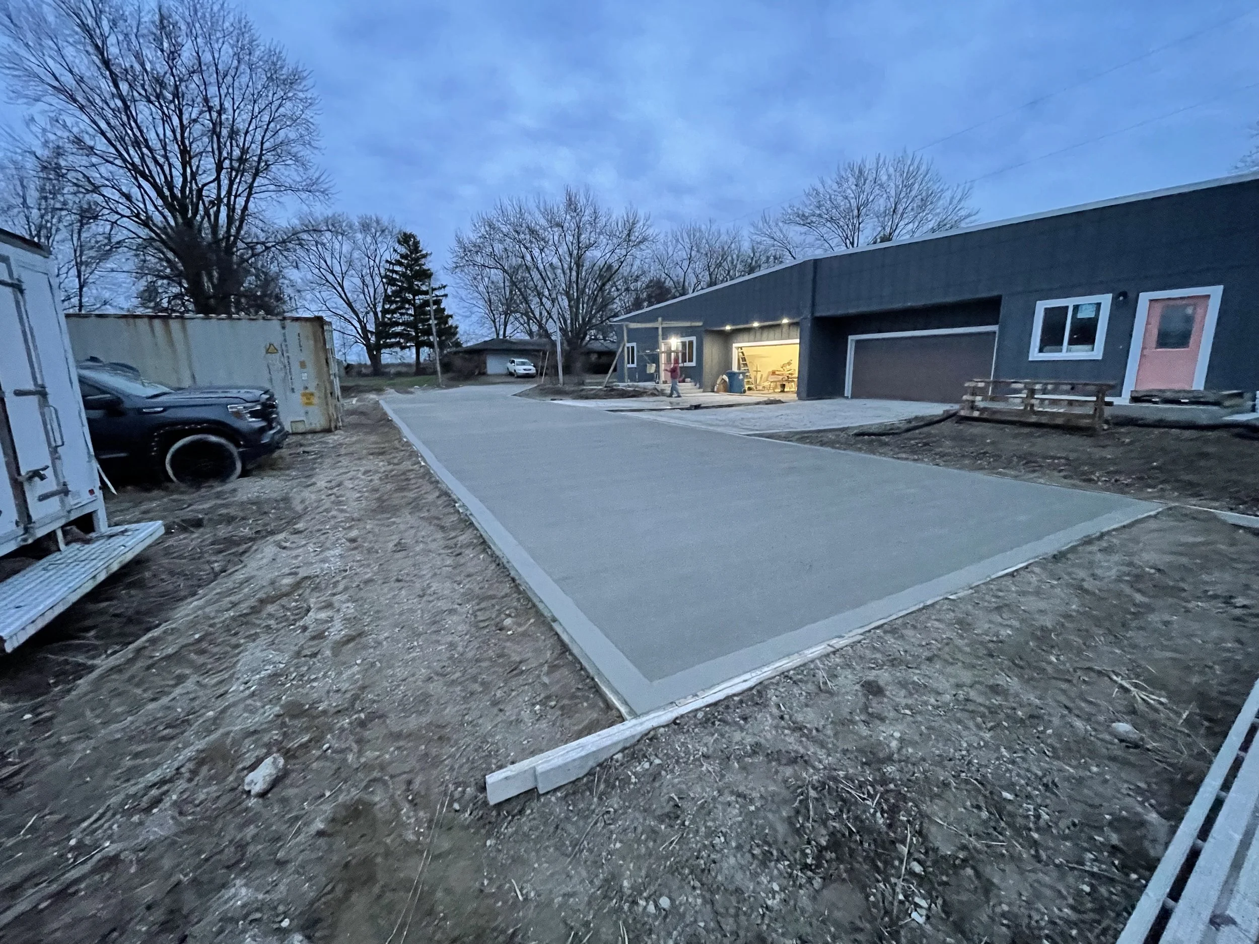 A newly poured concrete driveway in front of a modern home with lights on inside, surrounded by dirt and construction materials, with leafless trees in the background.