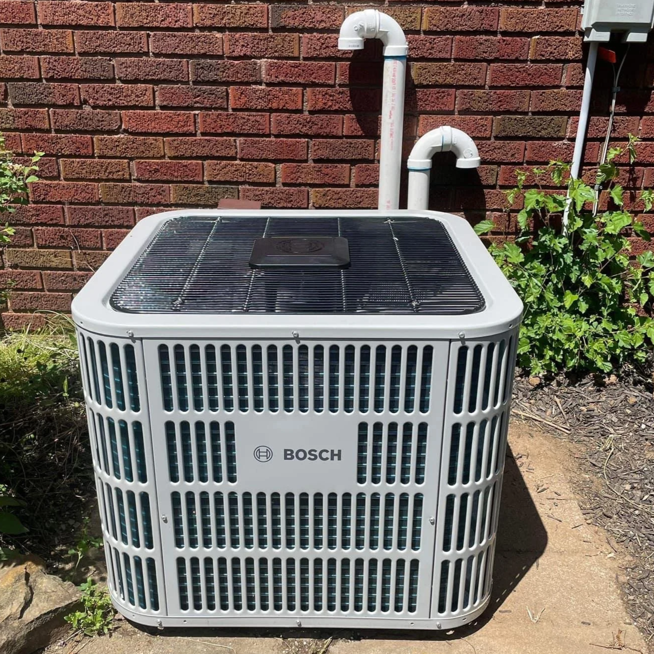 Outside view of a Bosch air conditioning unit with white protective grill, placed on a concrete surface near a red brick wall, with white PVC pipes and greenery in the background.