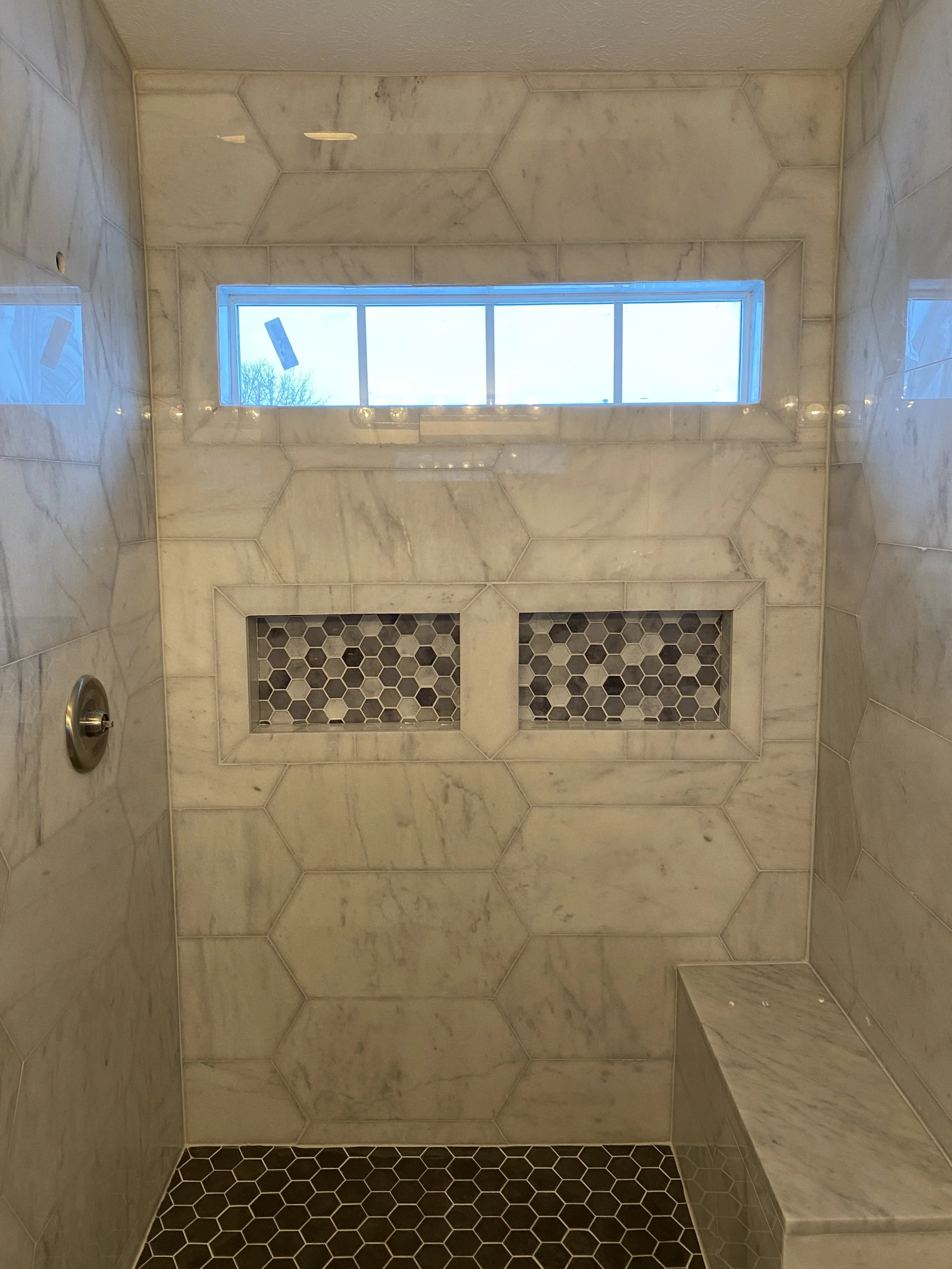 View of a tiled shower with marble hexagonal tiles on the walls, black hexagonal tiles on the floor, a long horizontal frosted window at the top, built-in marble shelves with hexagonal tiles, and a small marble bench.