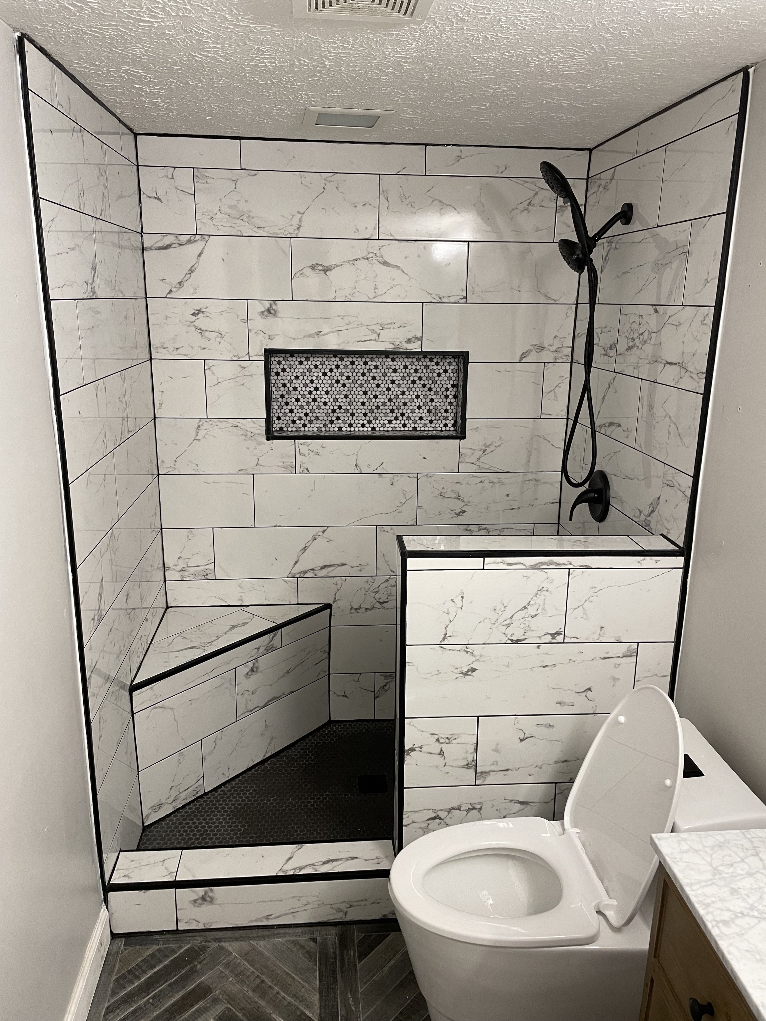 Bathroom with a walk-in shower featuring large white marble tiles, a small built-in bench, and a black showerhead. Toilet is in front of the shower, and part of a wooden vanity with a marble countertop is visible.