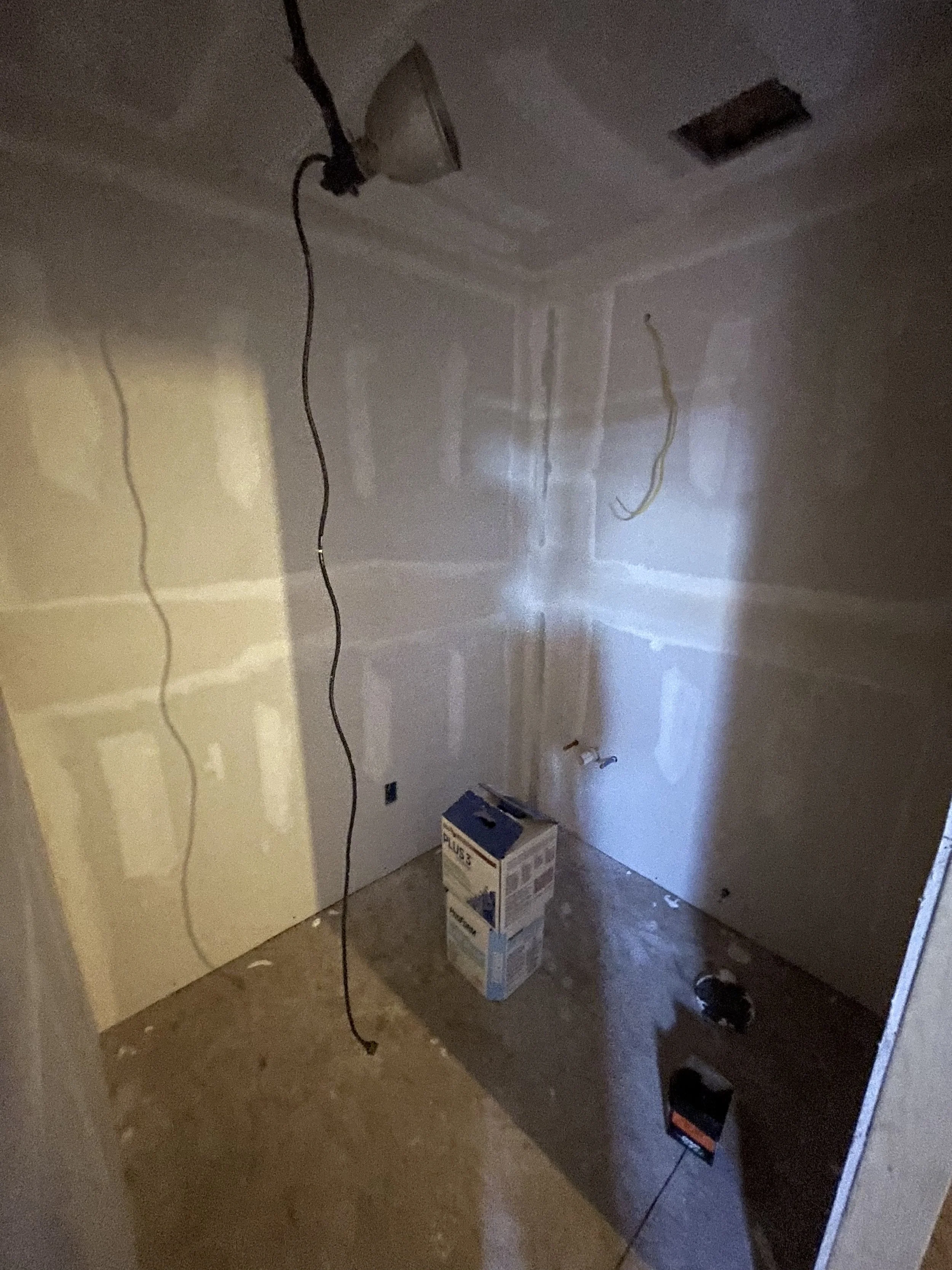 Unfinished room with drywall walls, construction materials, and electrical wiring hanging from the ceiling. A box and a small tool are on the floor.