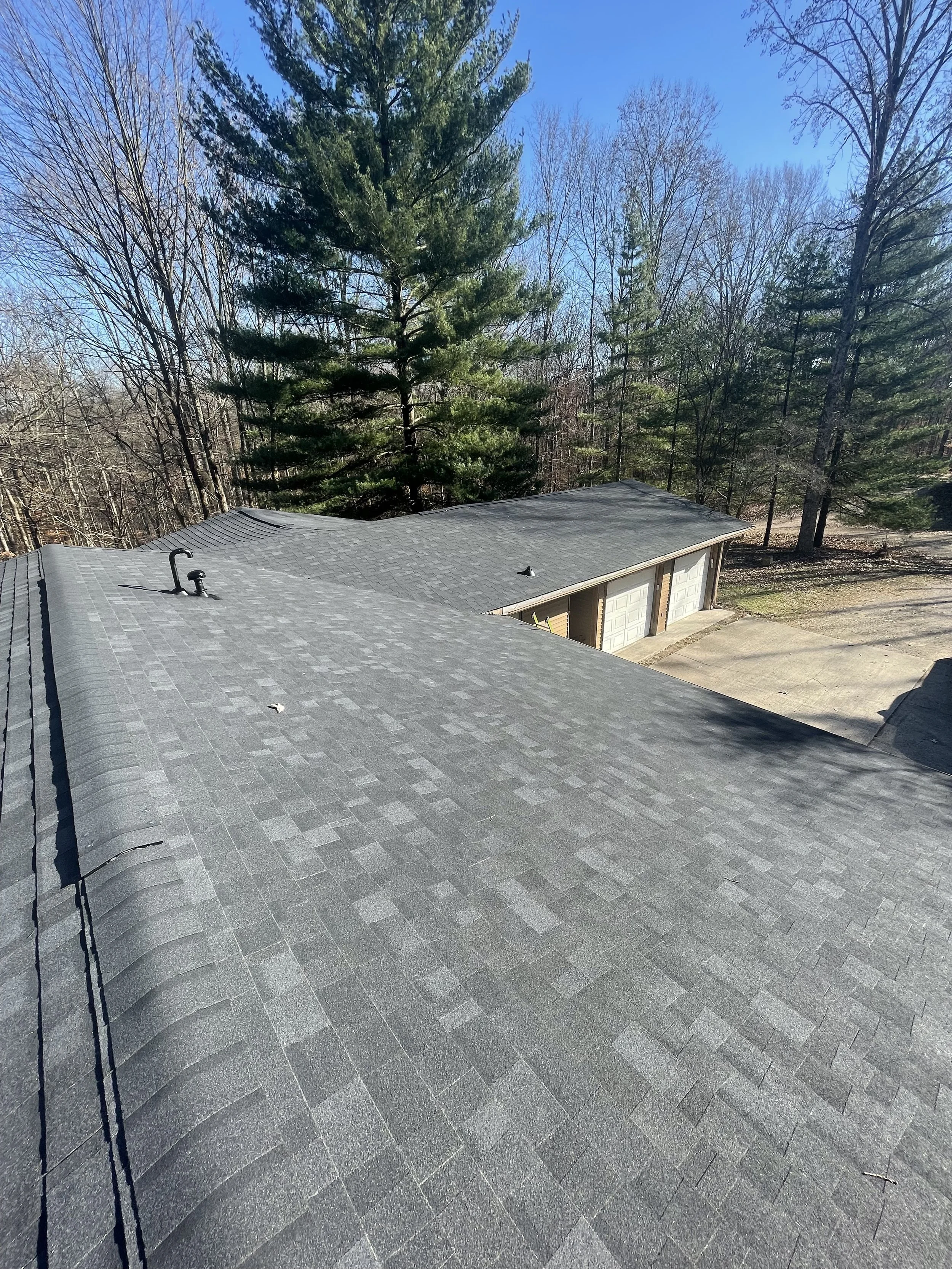 View of a black shingled roof of a house with a garage in the background, surrounded by tall trees, under a clear blue sky.