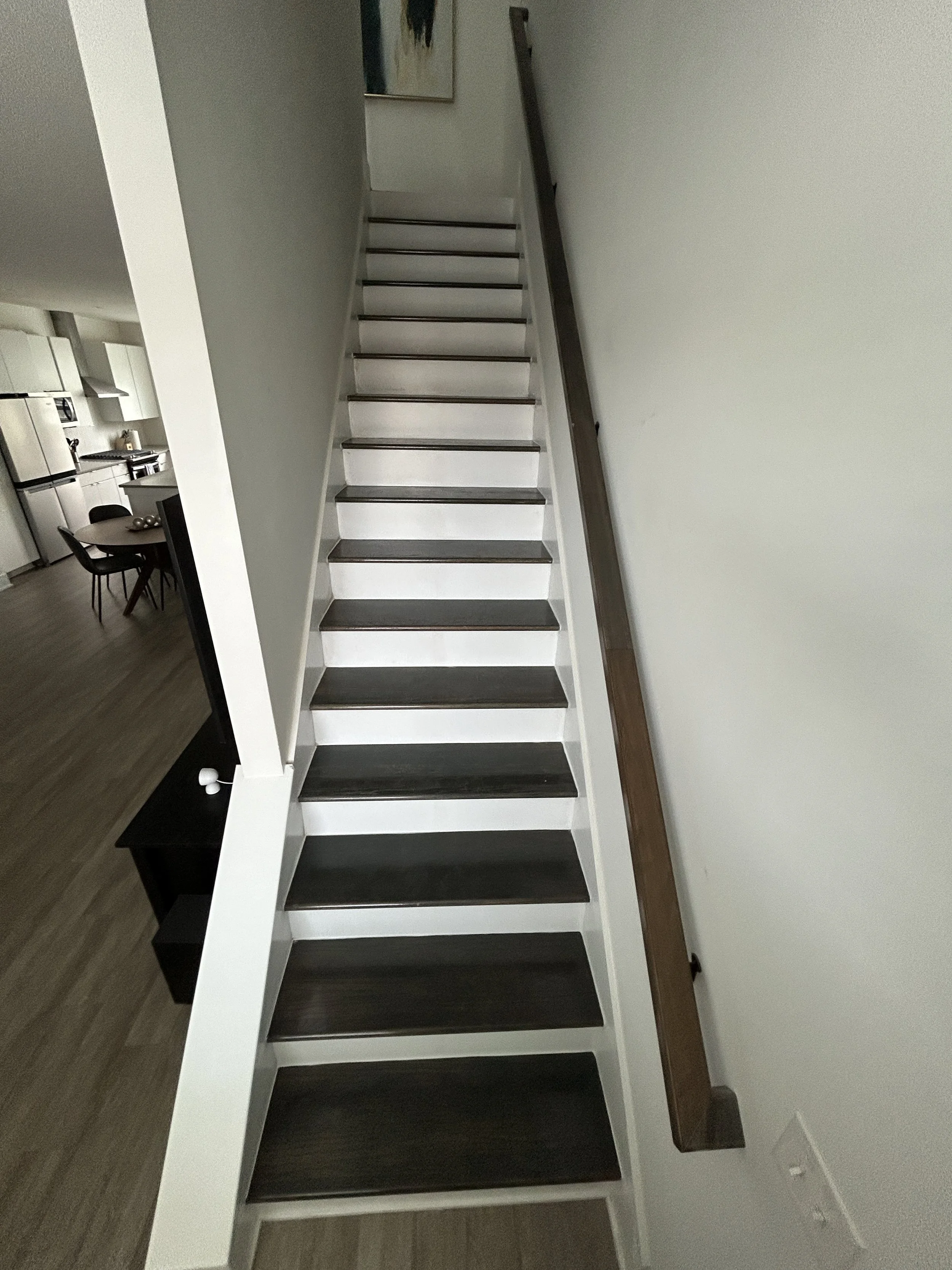 Interior staircase with dark wooden steps and white risers, dark wooden handrail on the right side, and a wall with a piece of artwork at the top. Part of a dining area with a table and chairs visible on the left.