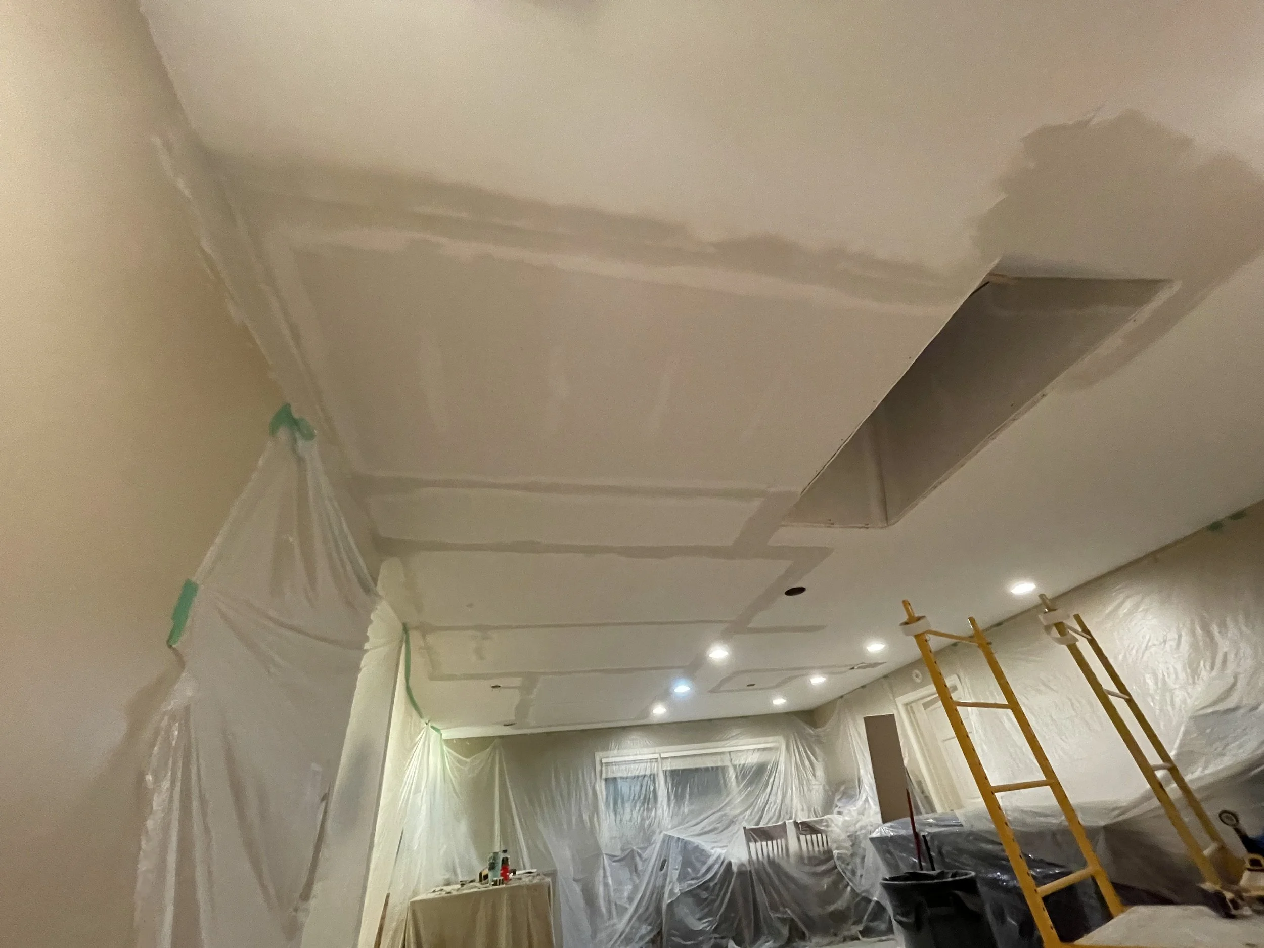 Ceiling being renovated with drywall and joint compound, with a ladder and plastic sheeting covering furniture and walls.
