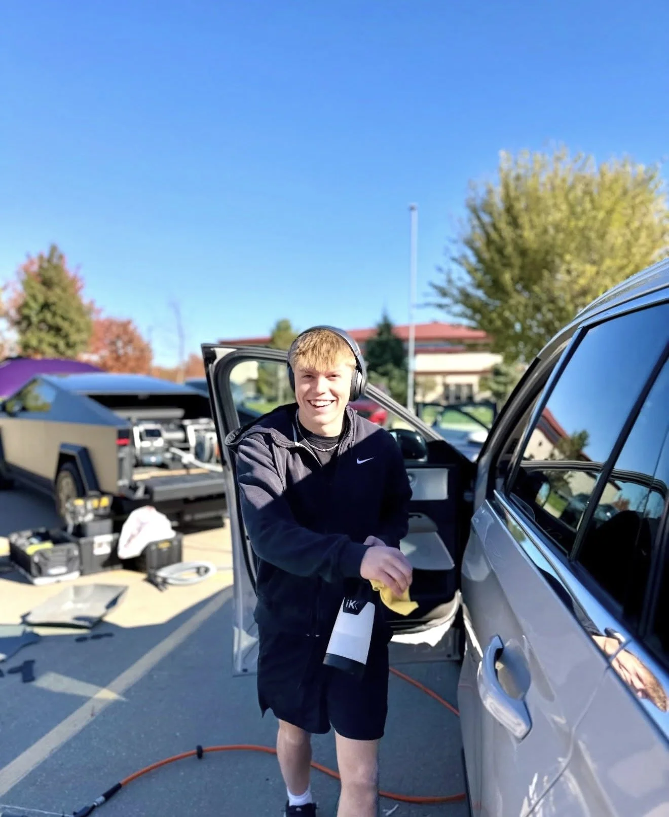 A smiling teenage boy with headphones washing a silver car in a parking lot on a sunny day.