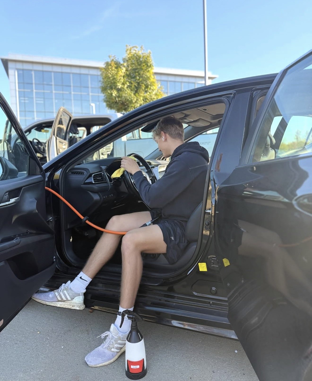 A young man sitting in the driver's seat of a black car, cleaning the interior with a spray bottle on the ground, during daytime with blue sky and modern glass building in the background.