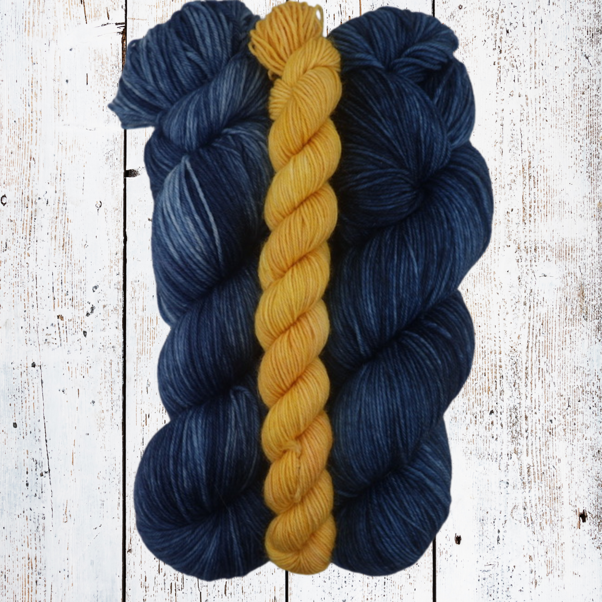 Two skeins of deep blue yarn with a yellow skein laid across them on a white wooden surface, highlighting the bold blue‑and‑yellow colour pairing and fibre softness.