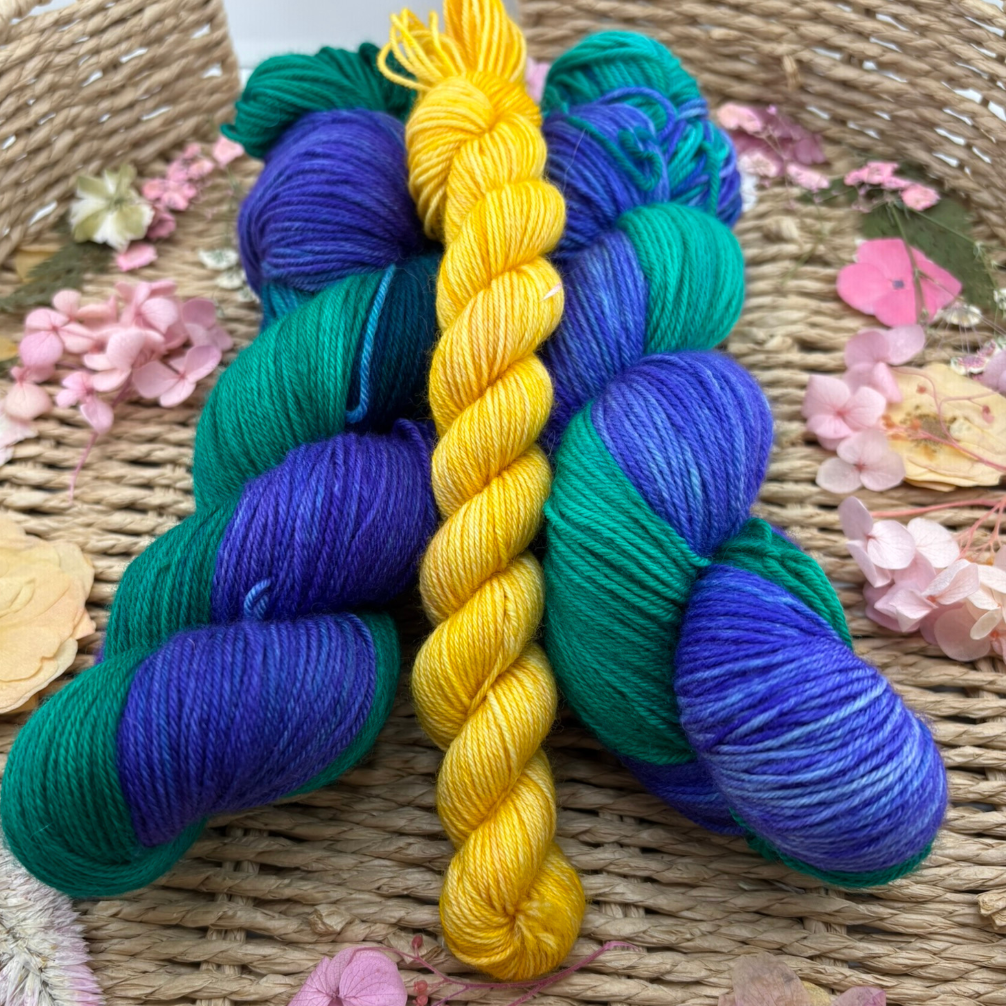 Yellow, blue and teal hand‑dyed yarn skeins styled together on a wicker surface with scattered pink petals, showing rich colour saturation and soft texture.