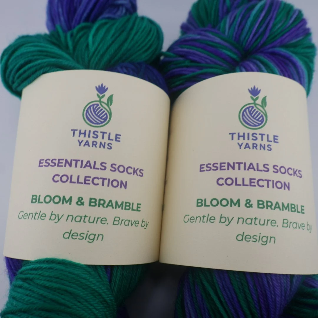 Two skeins of Thistle Yarns Essentials Socks Collection in Bloom & Bramble colorway with purple, green, and blue yarns.