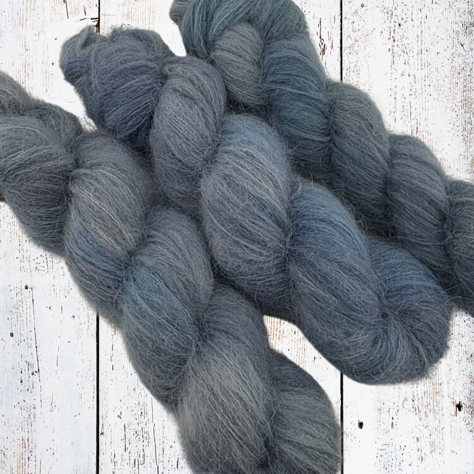 Blue‑grey alpaca yarn skeins with fluffy halo, hand‑dyed in small batches and styled on a rustic wooden surface.
