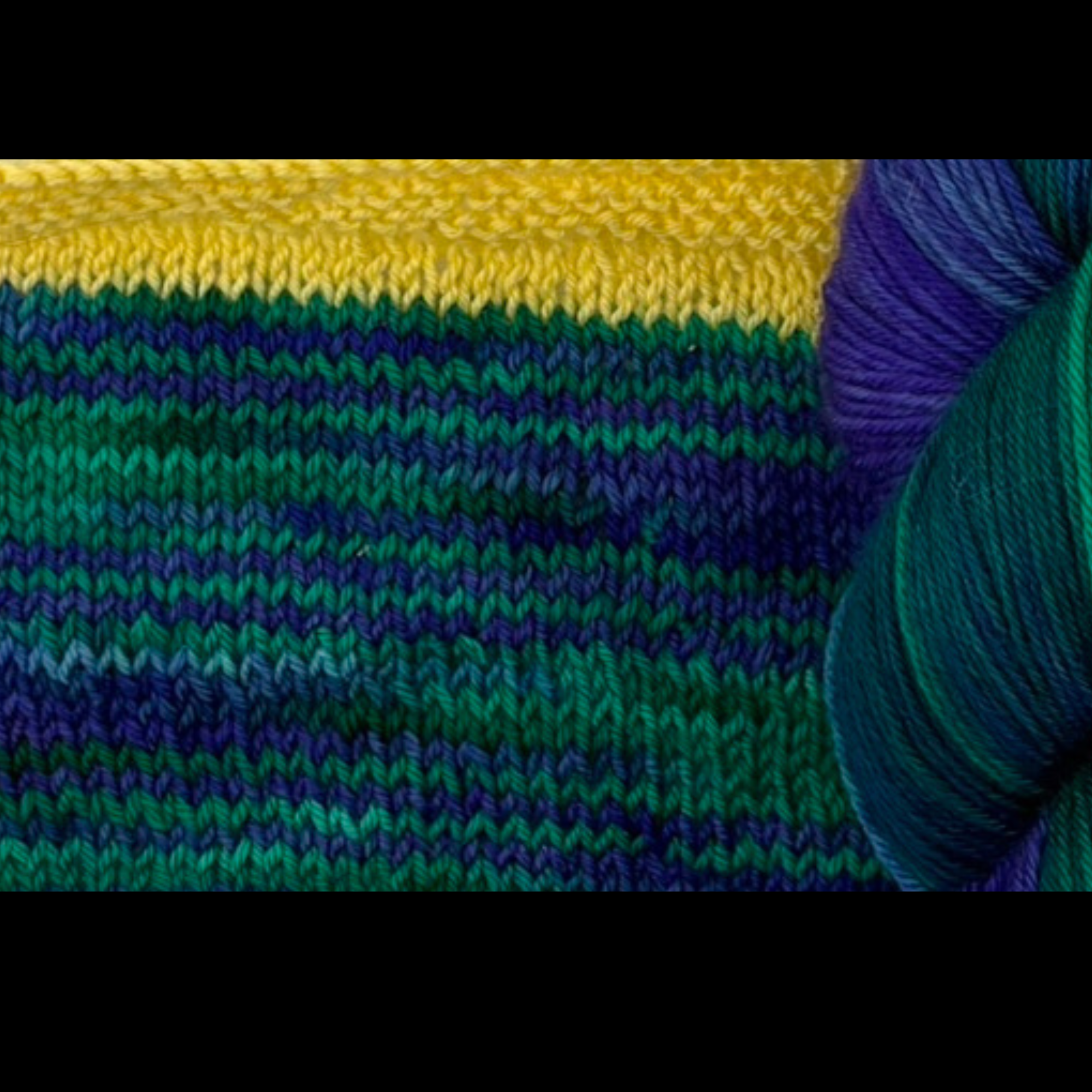 Close‑up of knitted fabric in horizontal stripes of yellow, green, blue and purple, showing how the hand‑dyed yarn blends and transitions when worked up.