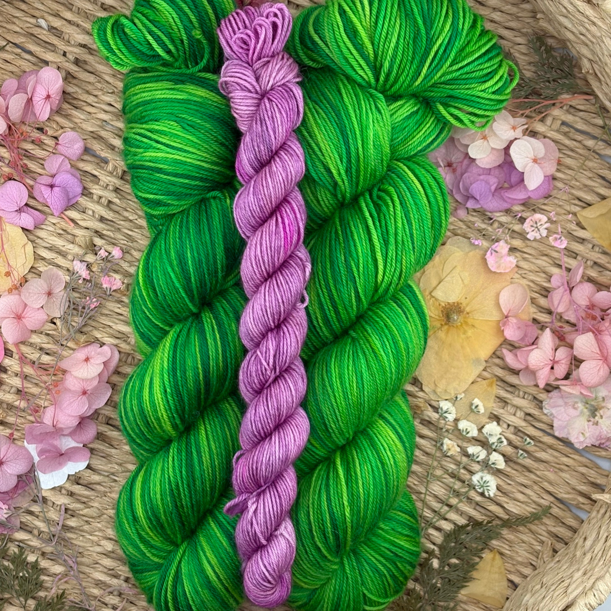Green and purple hand‑dyed yarn skeins displayed in a woven basket with dried petals, showing rich colour contrast and fibre texture.