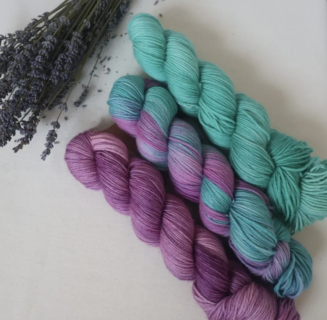 Multiple skeins of colorful yarn in shades of purple, pink, teal, and blue, along with a bunch of lavender flowers on a white background.