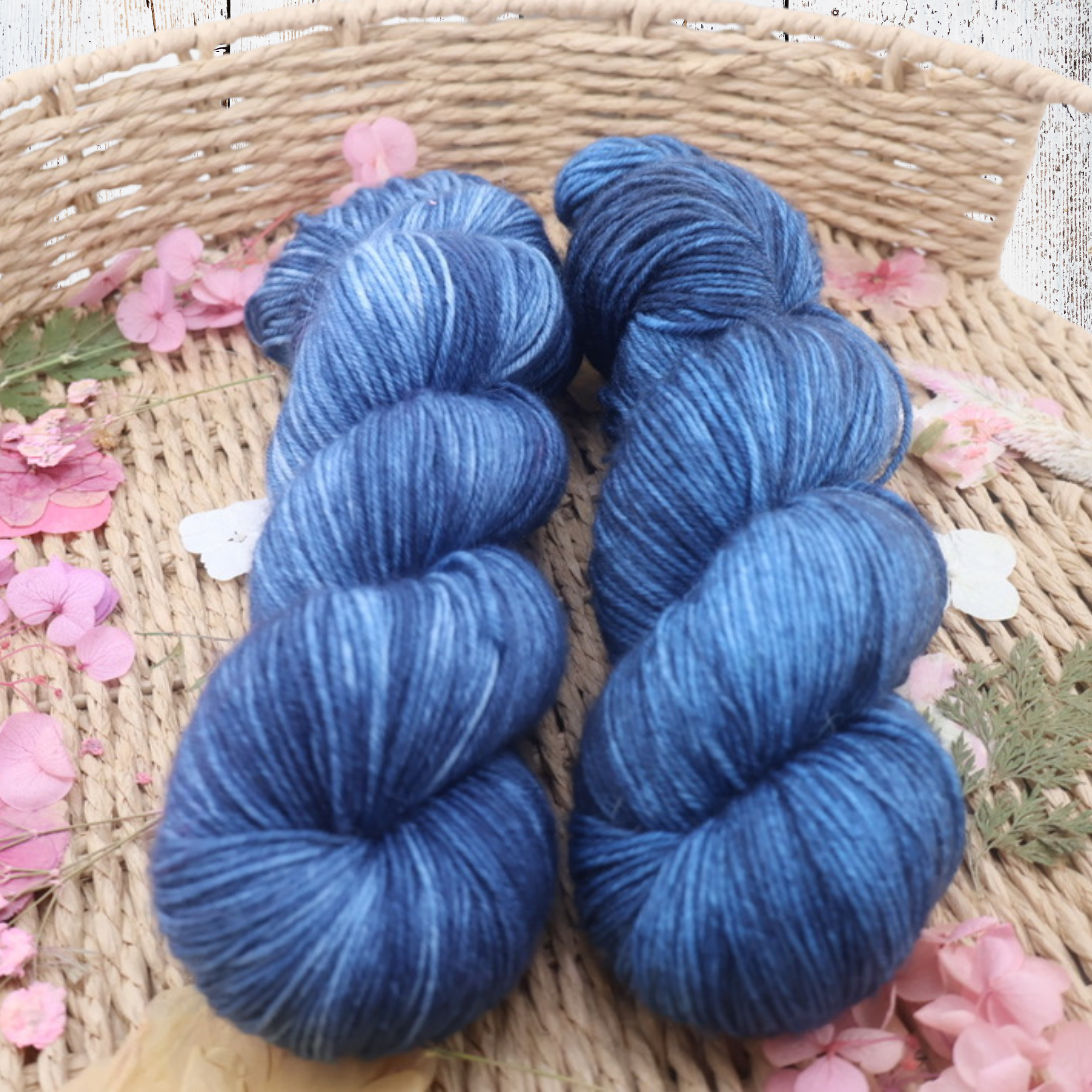 Two skeins of deep blue hand‑dyed yarn in a woven basket with pink flower petals on a white wooden surface, showing soft texture and rich colour saturation.