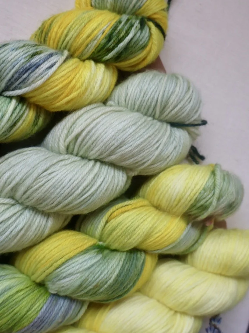 Close‑up of artisan‑dyed Merino DK skeins showing variegated greys, greens, and bright yellows.