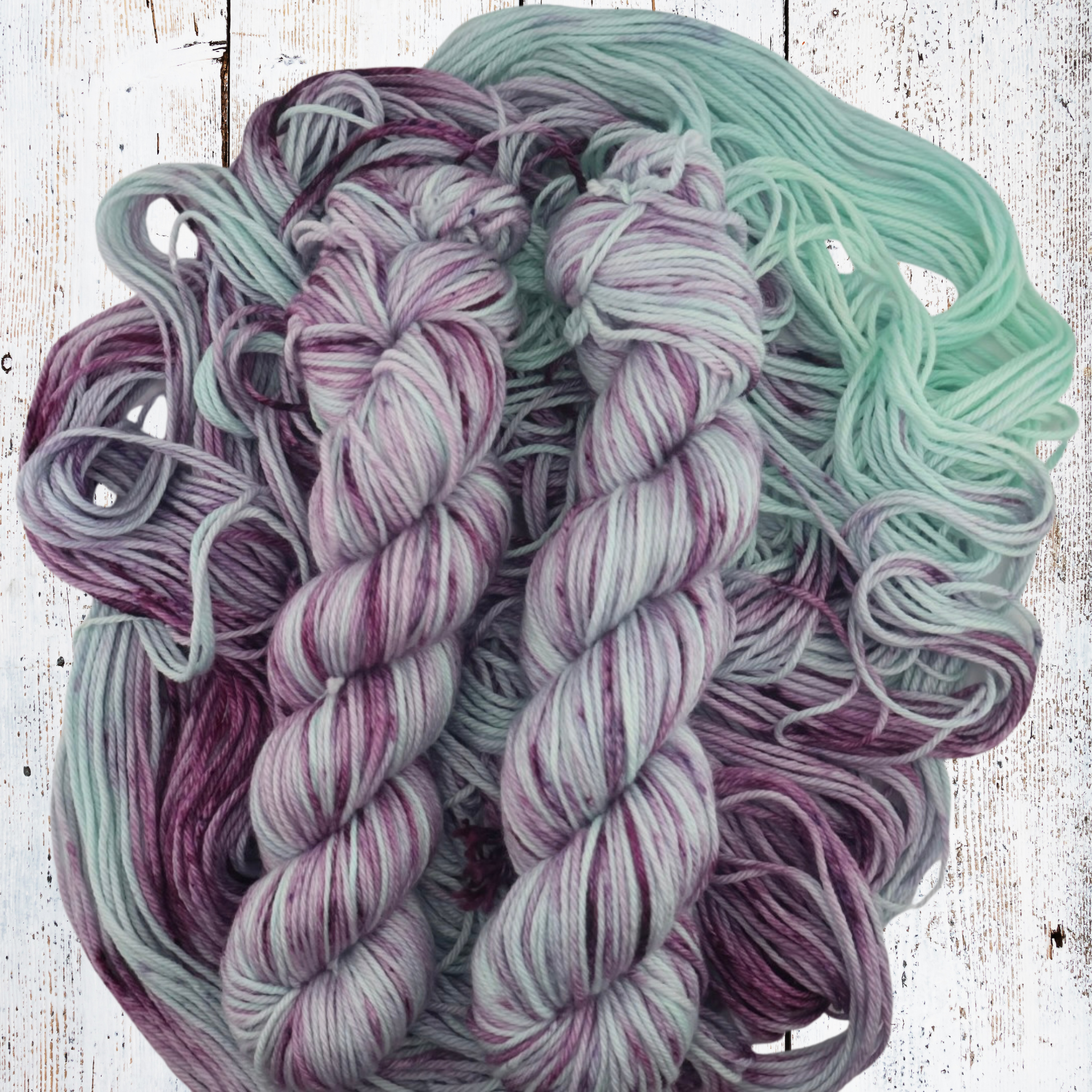 Three purple and grey yarn skeins displayed on a white wooden surface surrounded by loose strands of matching fibre